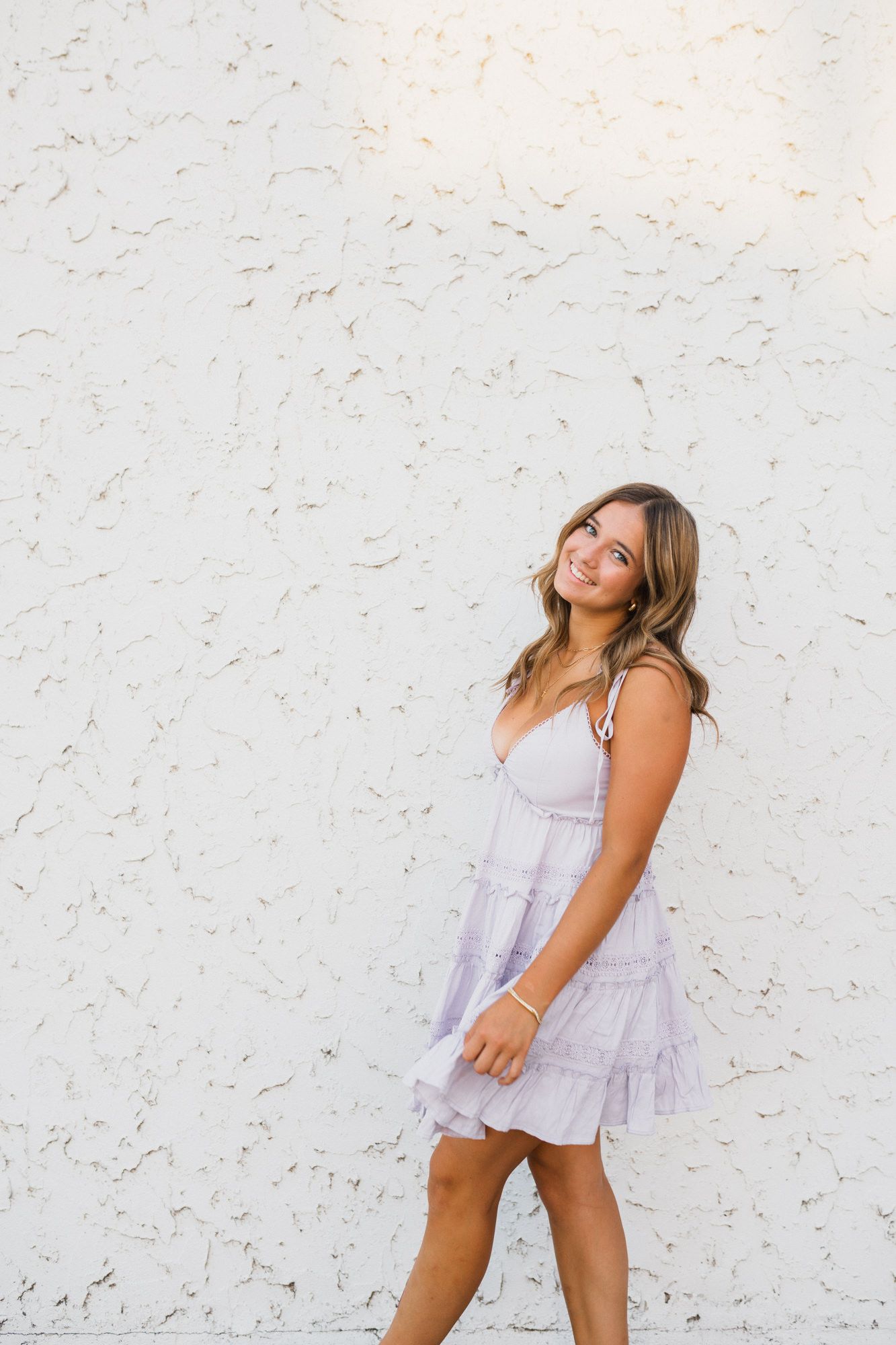 Young woman in lavender mini dress smiling against white textured wall.