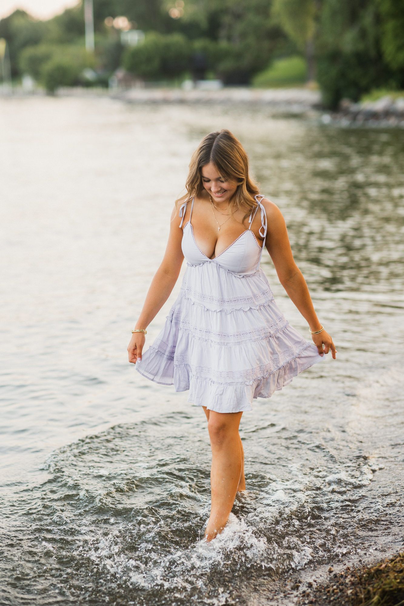 Woman in white sundress wading in shallow water near shore, smiling and enjoying a warm summer day.