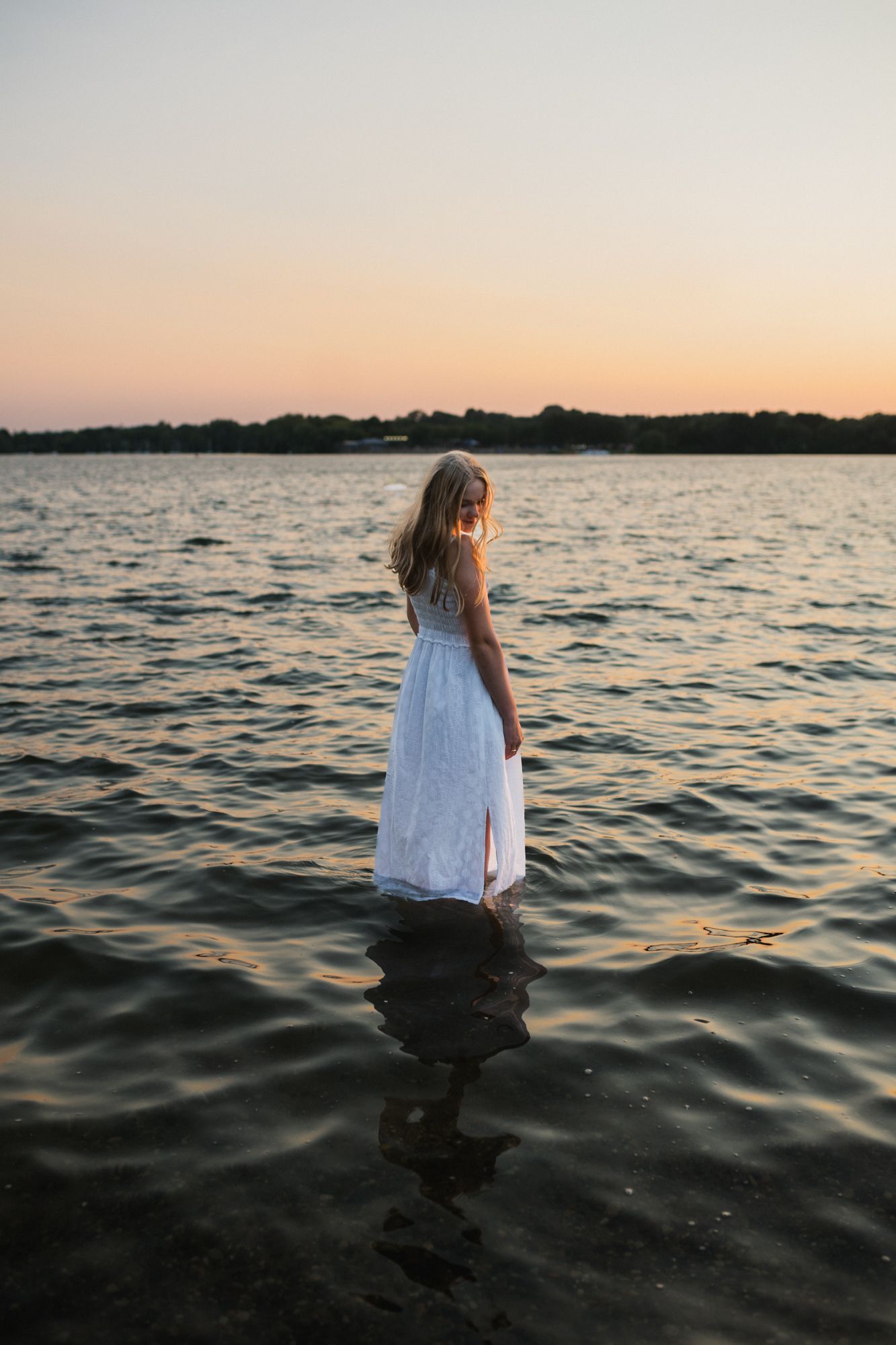 Girl in white dress standing in calm lake water at golden sunset with treeline silhouette in background.