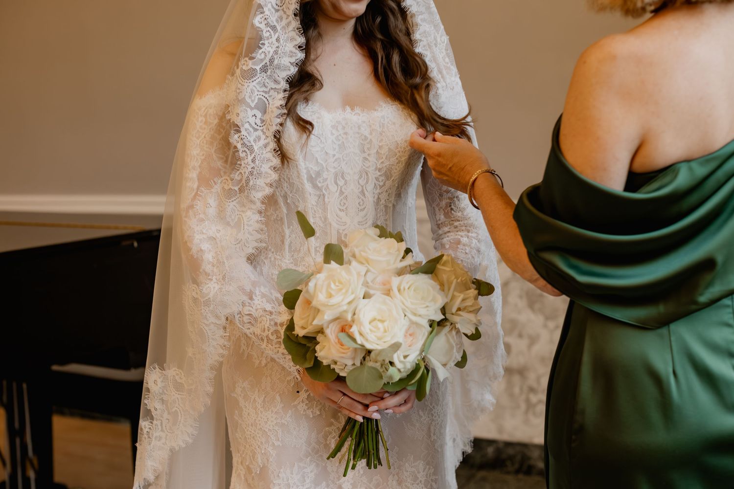 Bride in lace wedding gown and veil holds white rose bouquet while bridesmaid in green dress assists her.