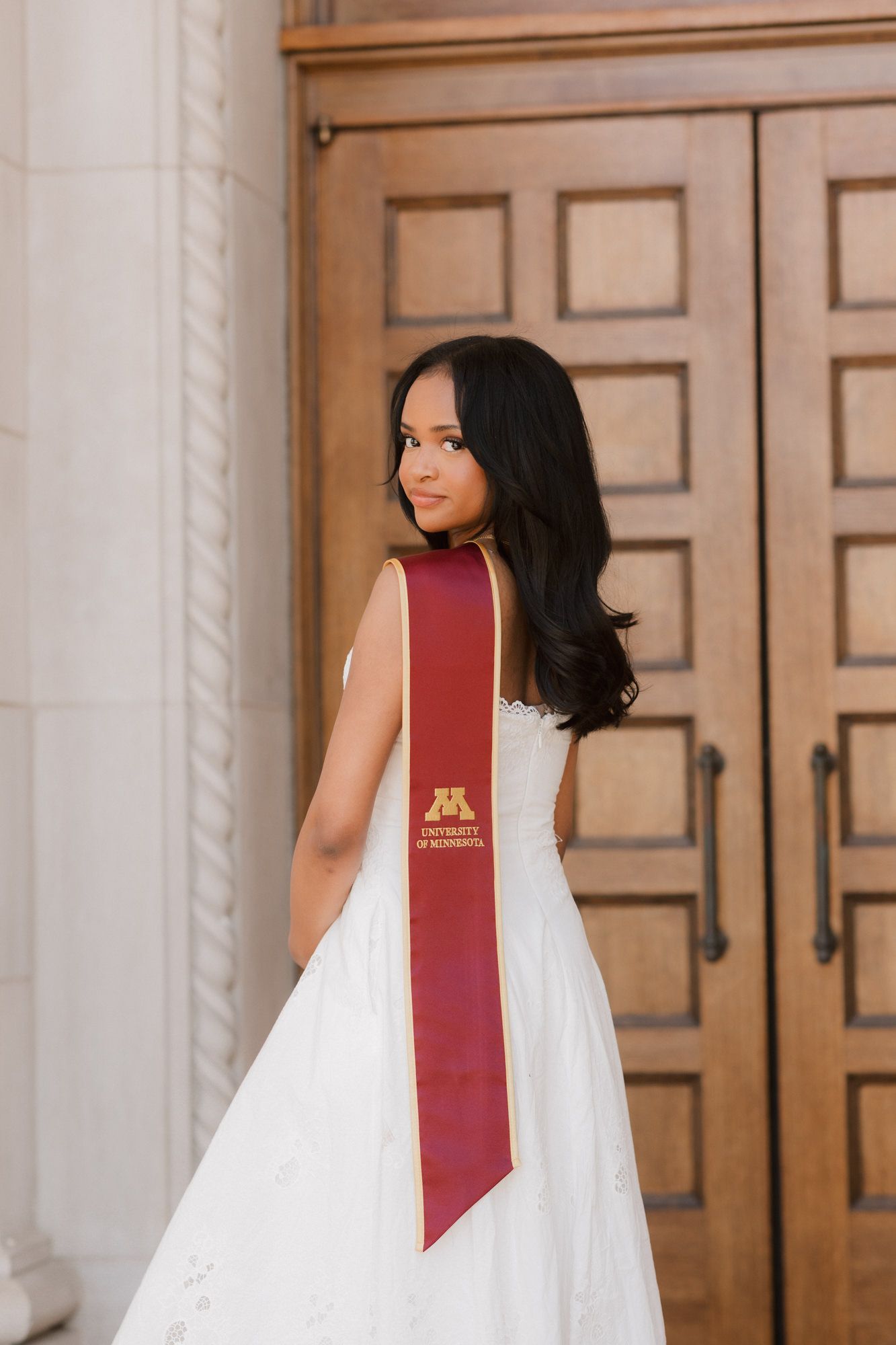 Graduate in white dress with University of Minnesota maroon sash poses before wooden doors.
