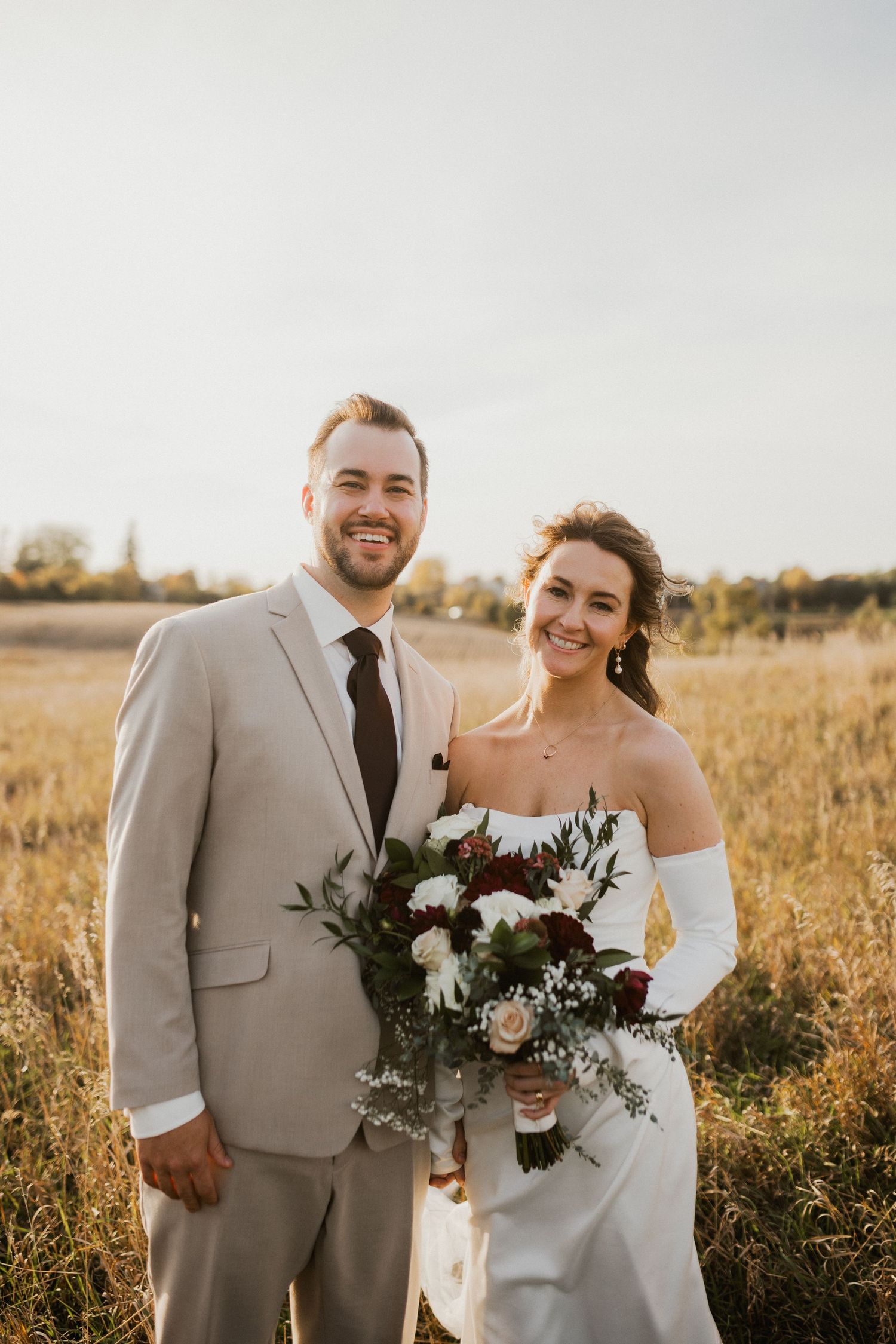 Smiling bride in a white strapless gown and groom in a tan suit hold a burgundy and white bouquet in a golden field.