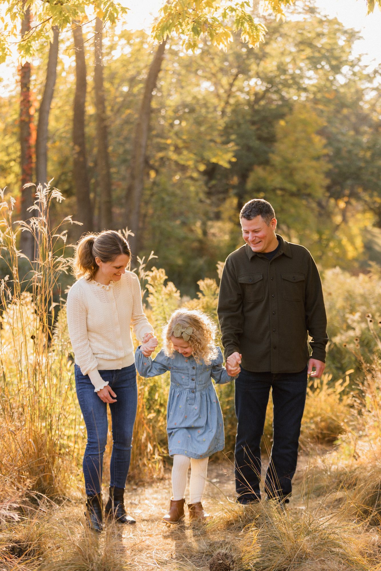 Family of three walking hand-in-hand on a sunlit autumn trail surrounded by golden grasses and trees.