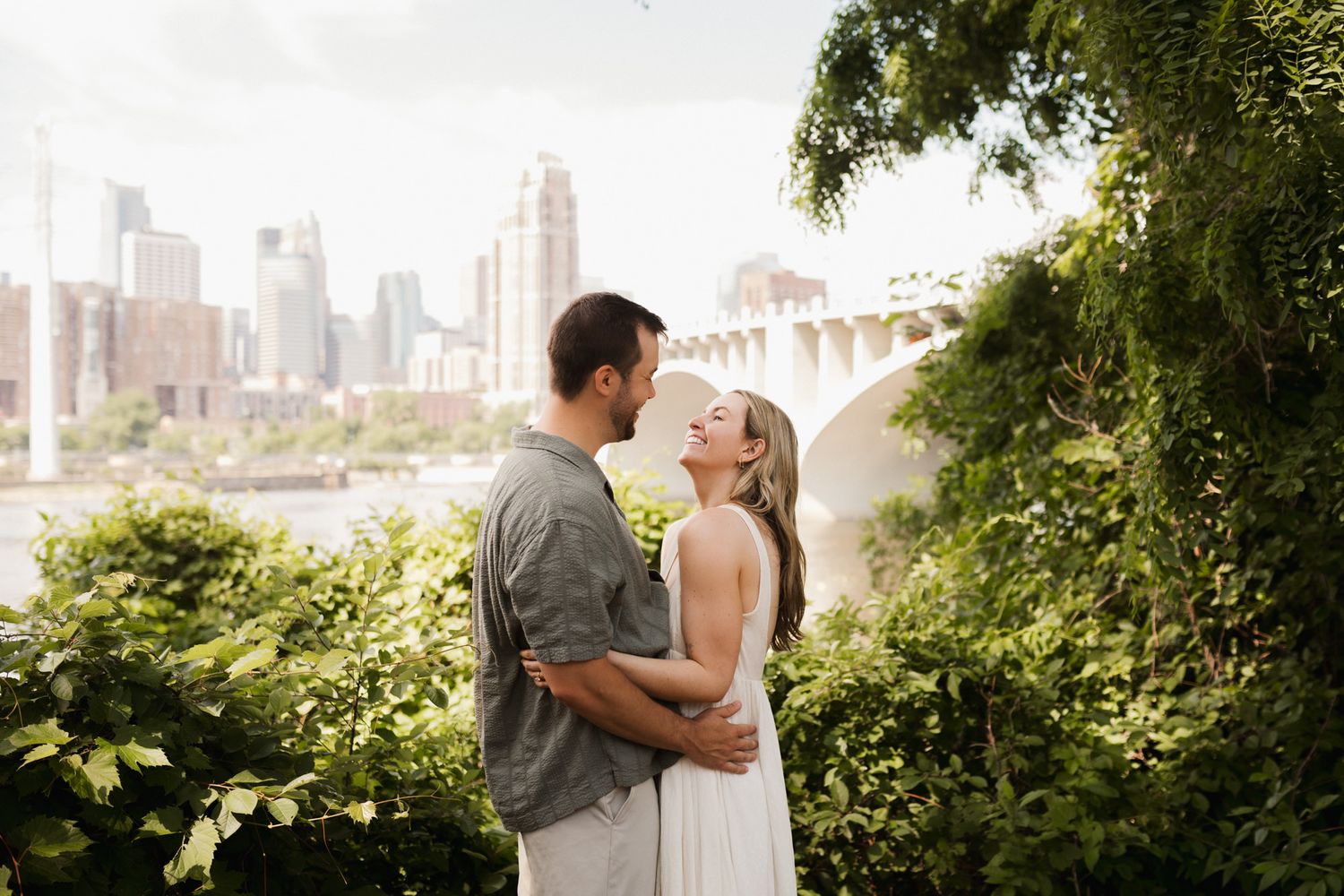 Couple embracing and almost kissing outdoors with lush greenery and city skyline in the background.