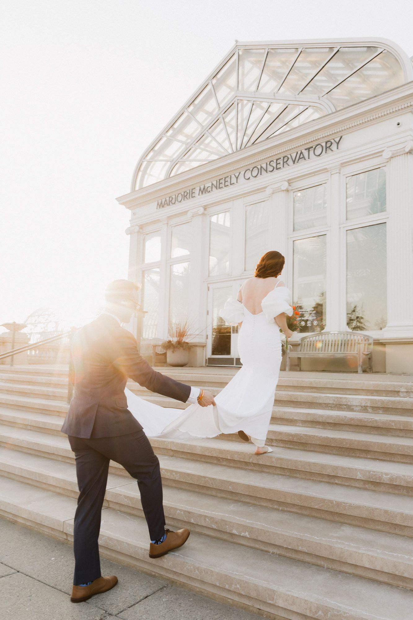 Bride and groom walking up steps to Marjorie McNeely Conservatory in golden sunlight, bride in backless white gown.