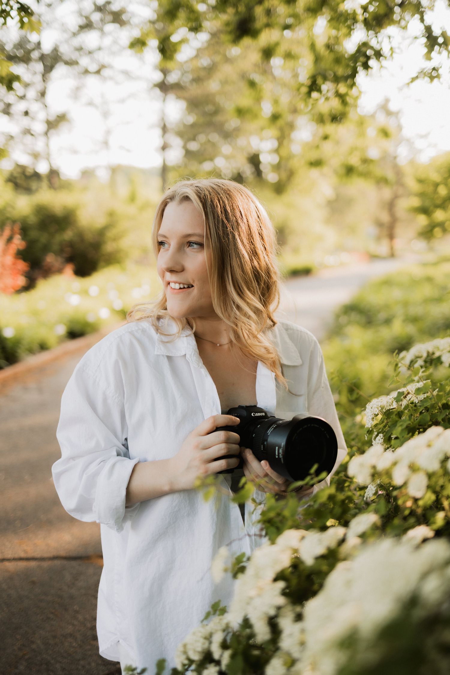 Smiling blonde woman holding a DSLR camera outdoors in a lush green garden wearing a white linen shirt.
