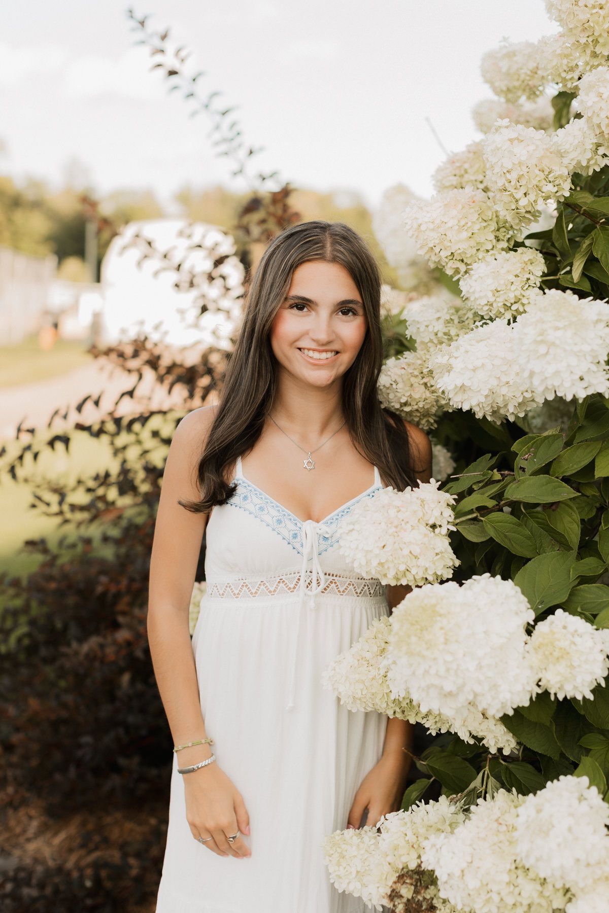Smiling brunette teen in white lace dress posing beside blooming white hydrangea bushes outdoors.