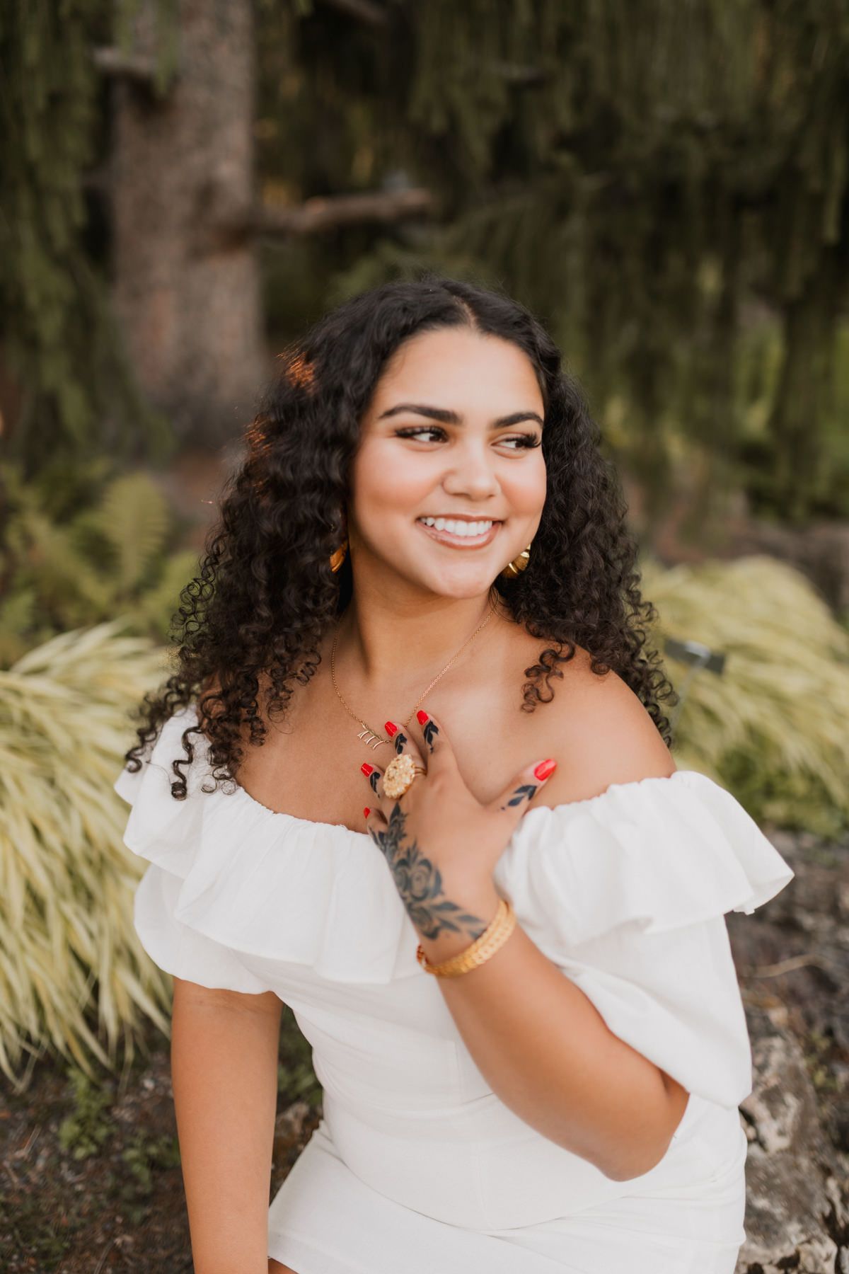 Smiling young woman with curly hair wearing a white off-shoulder dress, gold jewelry, and red nails in a garden setting.