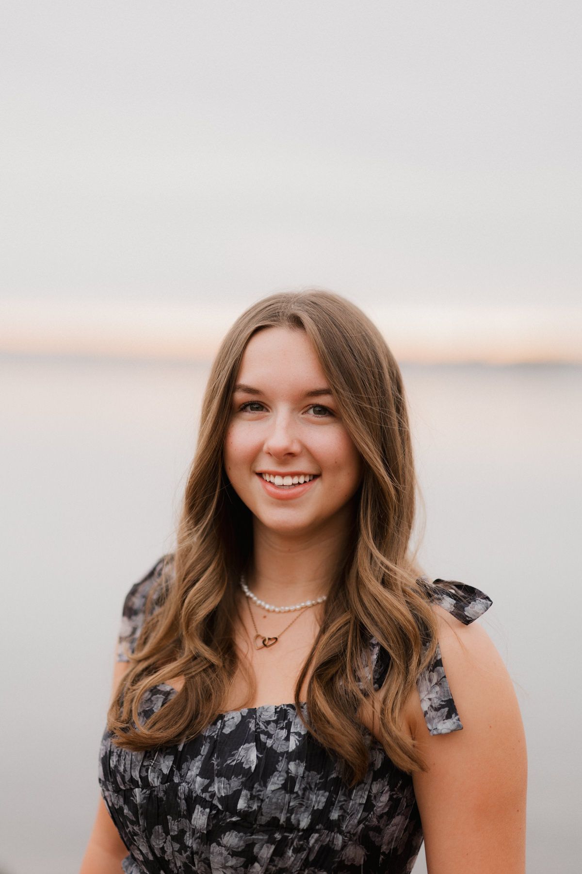 Smiling young woman with wavy blonde hair wearing a floral dress and pearl necklace poses by a calm waterfront.