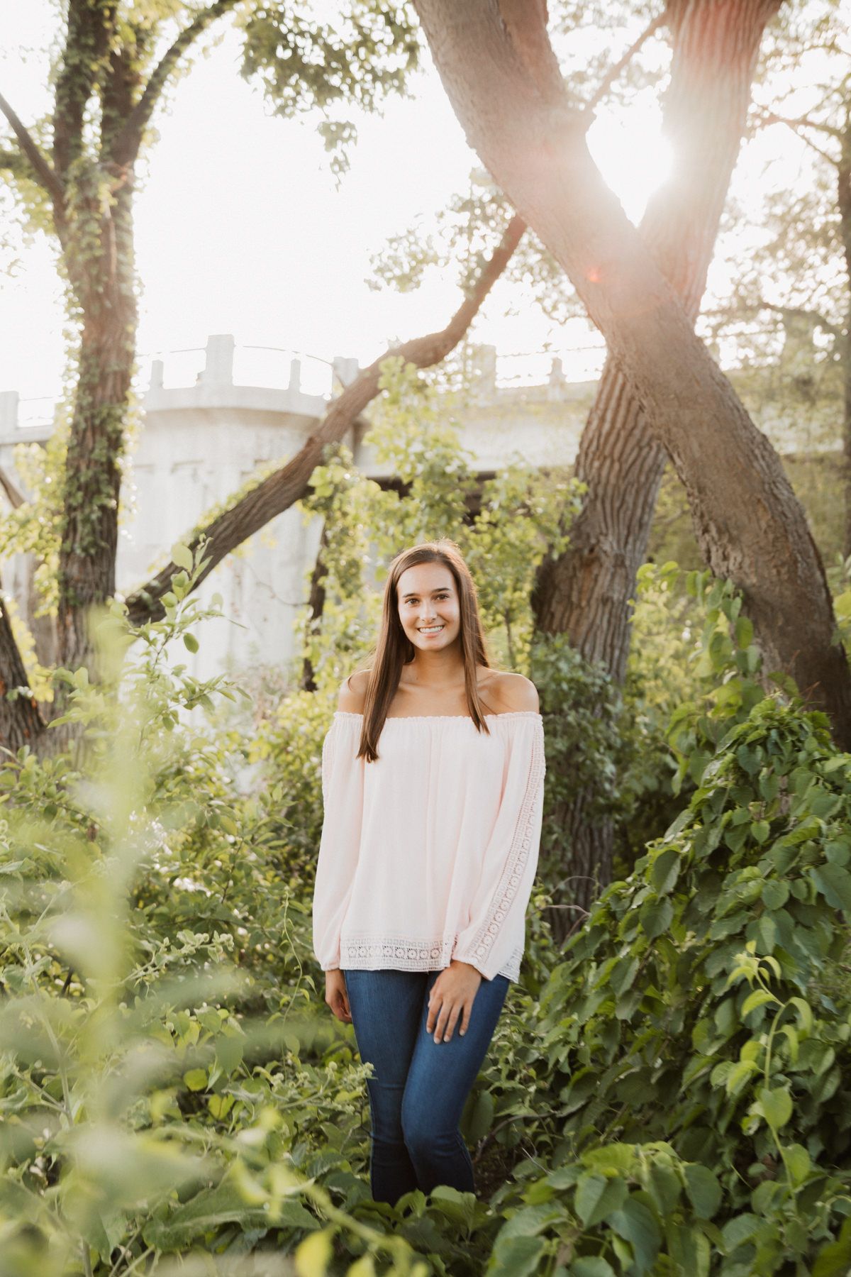 Young woman in off-shoulder pink top and jeans smiling among lush green foliage with sunlight filtering through trees.