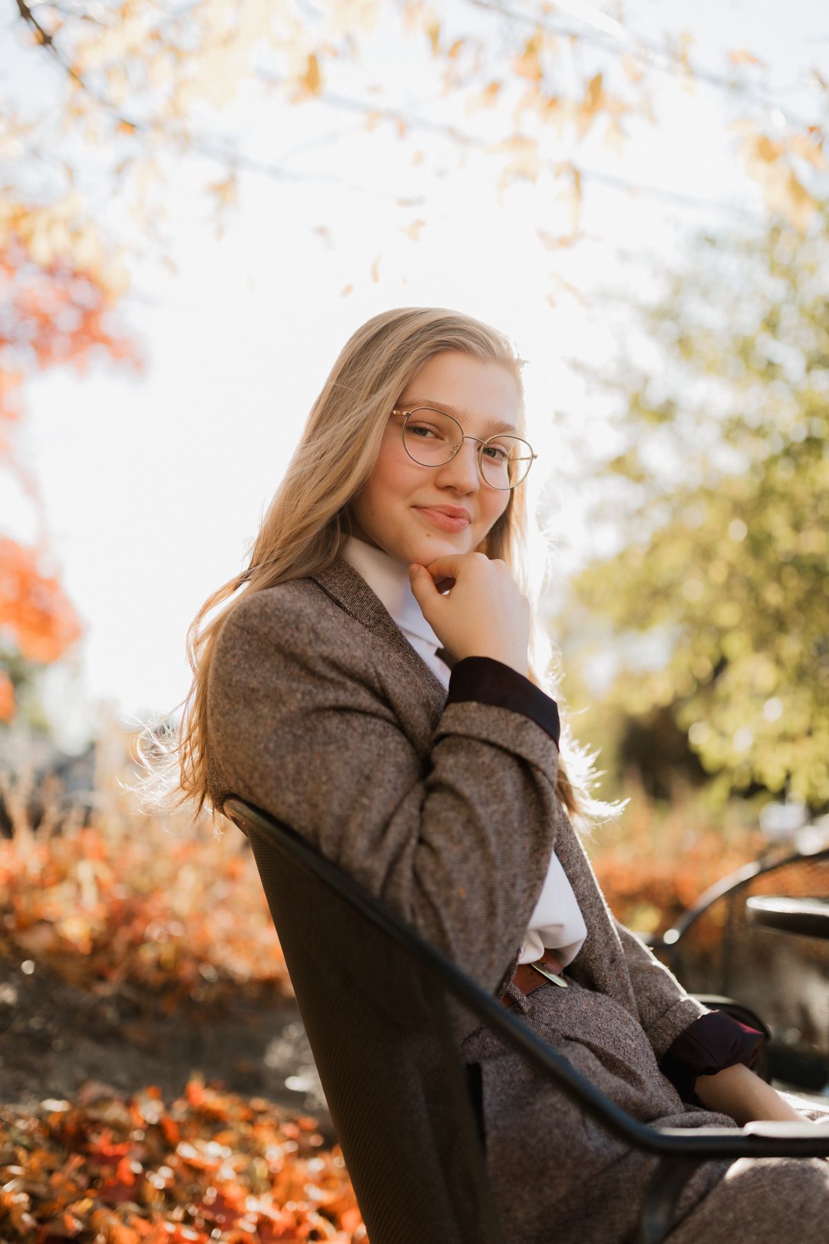 Blonde teen girl in gray coat and glasses sits on bench surrounded by autumn foliage.