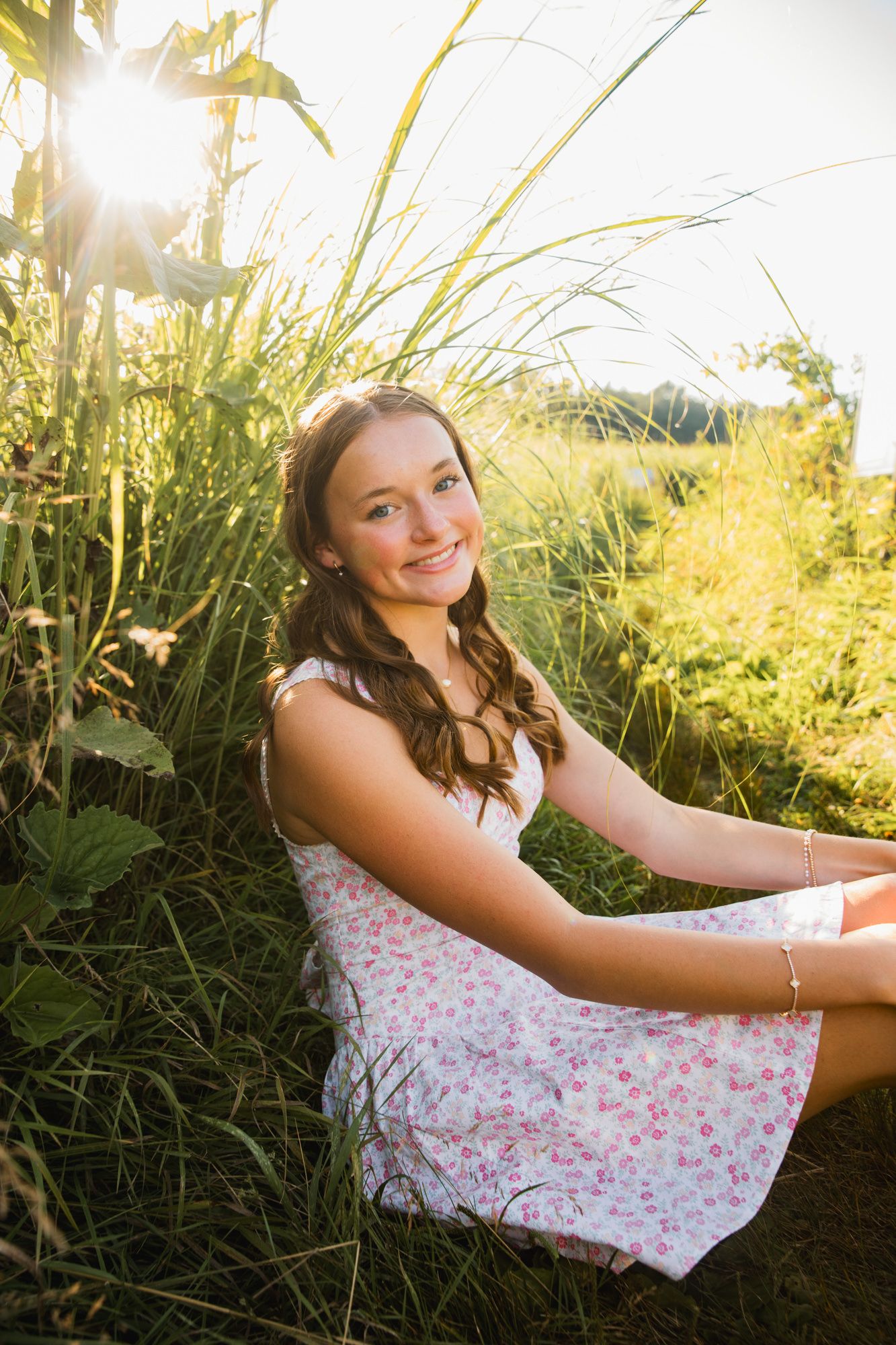 Young woman in floral dress sitting in tall grass, smiling in golden sunlight during outdoor portrait session.