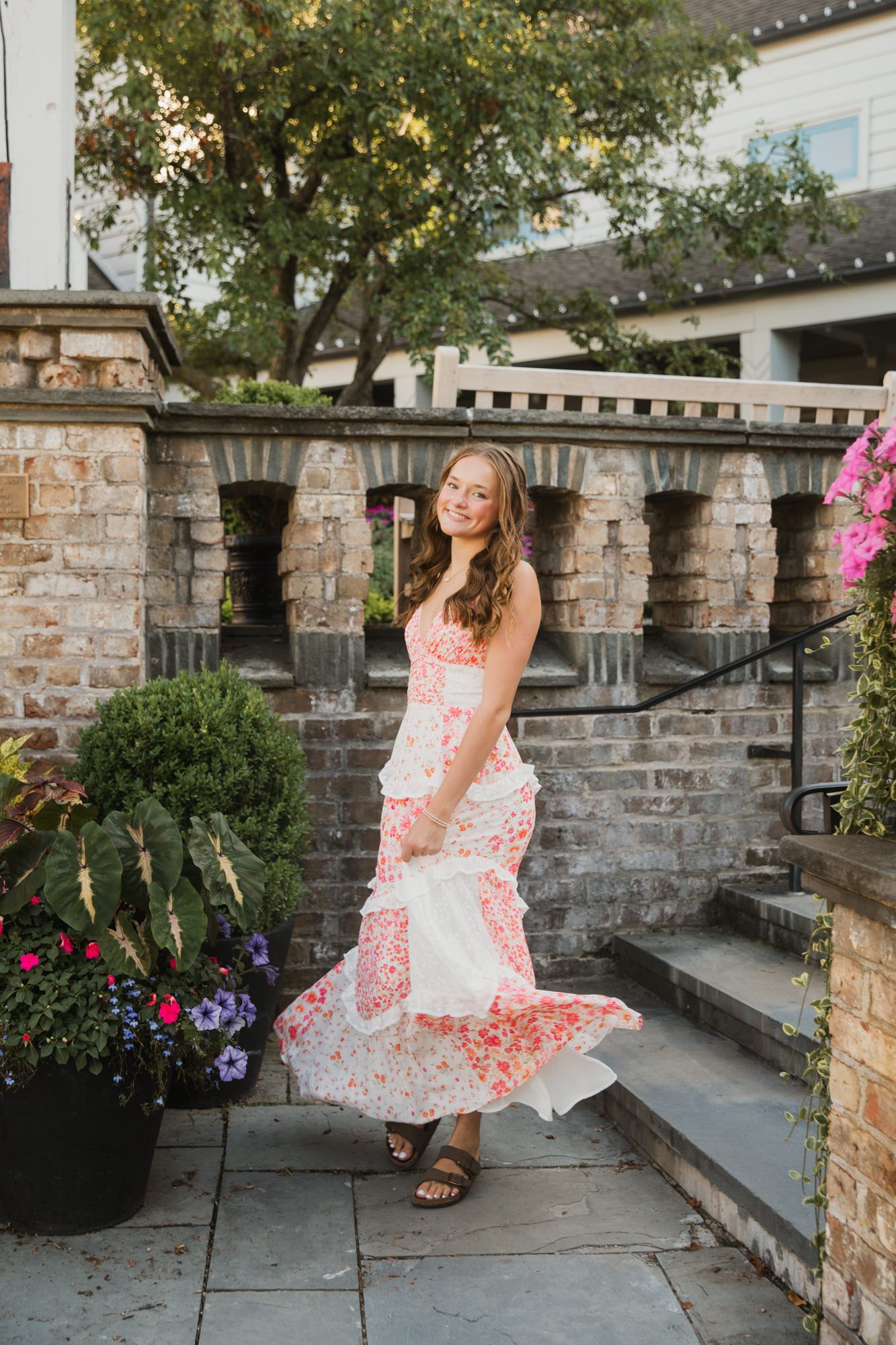 Young woman twirling in a floral maxi dress on a stone patio surrounded by lush greenery and flowers.