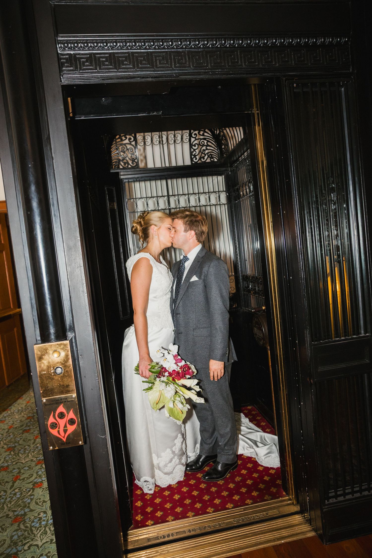 Bride and groom sharing a kiss inside a vintage ornate elevator holding a burgundy and white bridal bouquet.