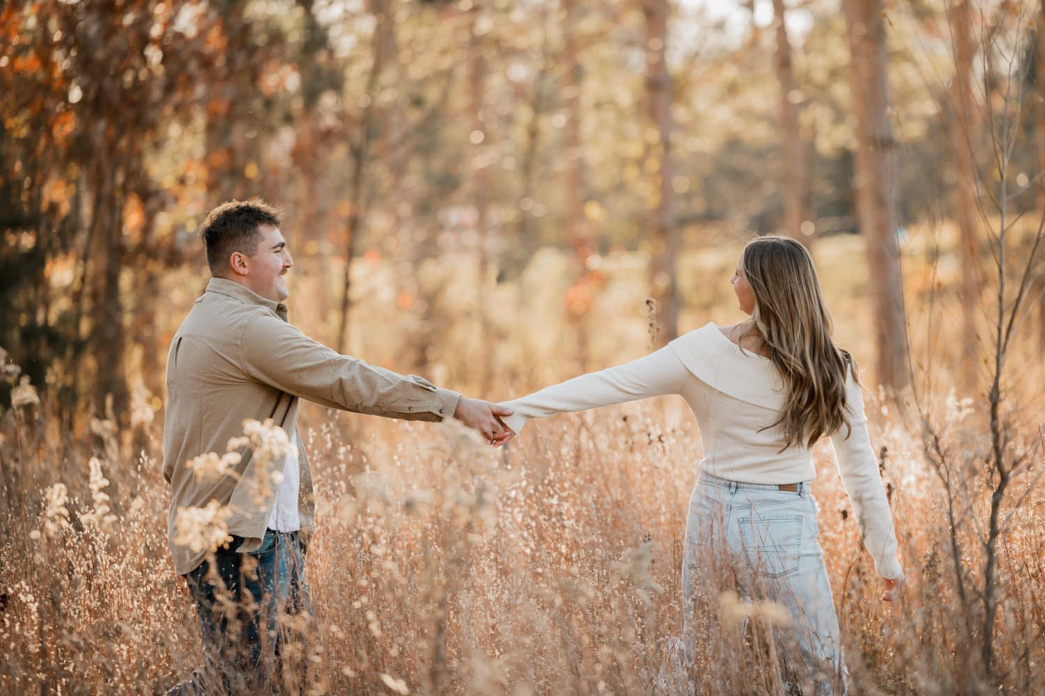 Two people playfully interact in a field of tall grass surrounded by autumn trees.