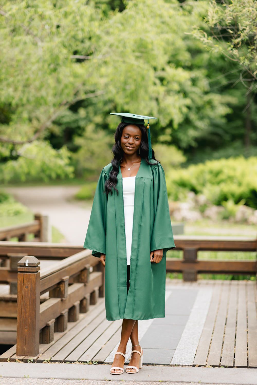 Graduate in emerald green cap and gown stands on wooden bridge pathway in garden setting.