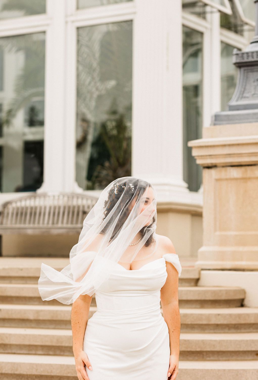 Bride in flowing white dress poses on grand stone steps of a conservatory.