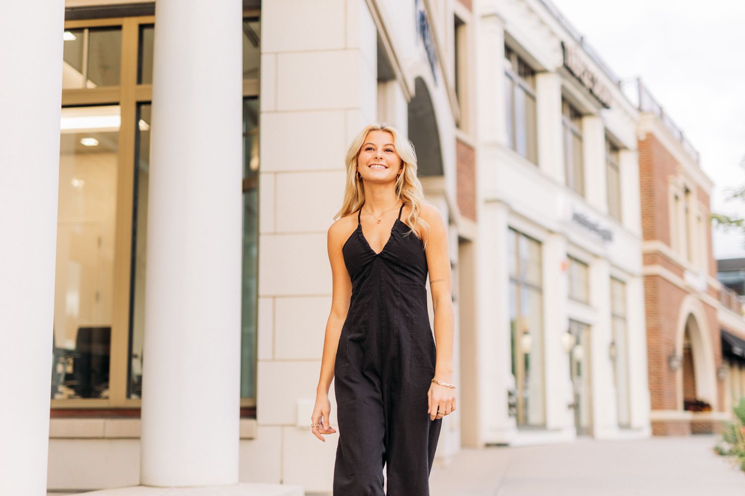 Woman in black sleeveless dress and boots stands elegantly against white stone wall with columns.
