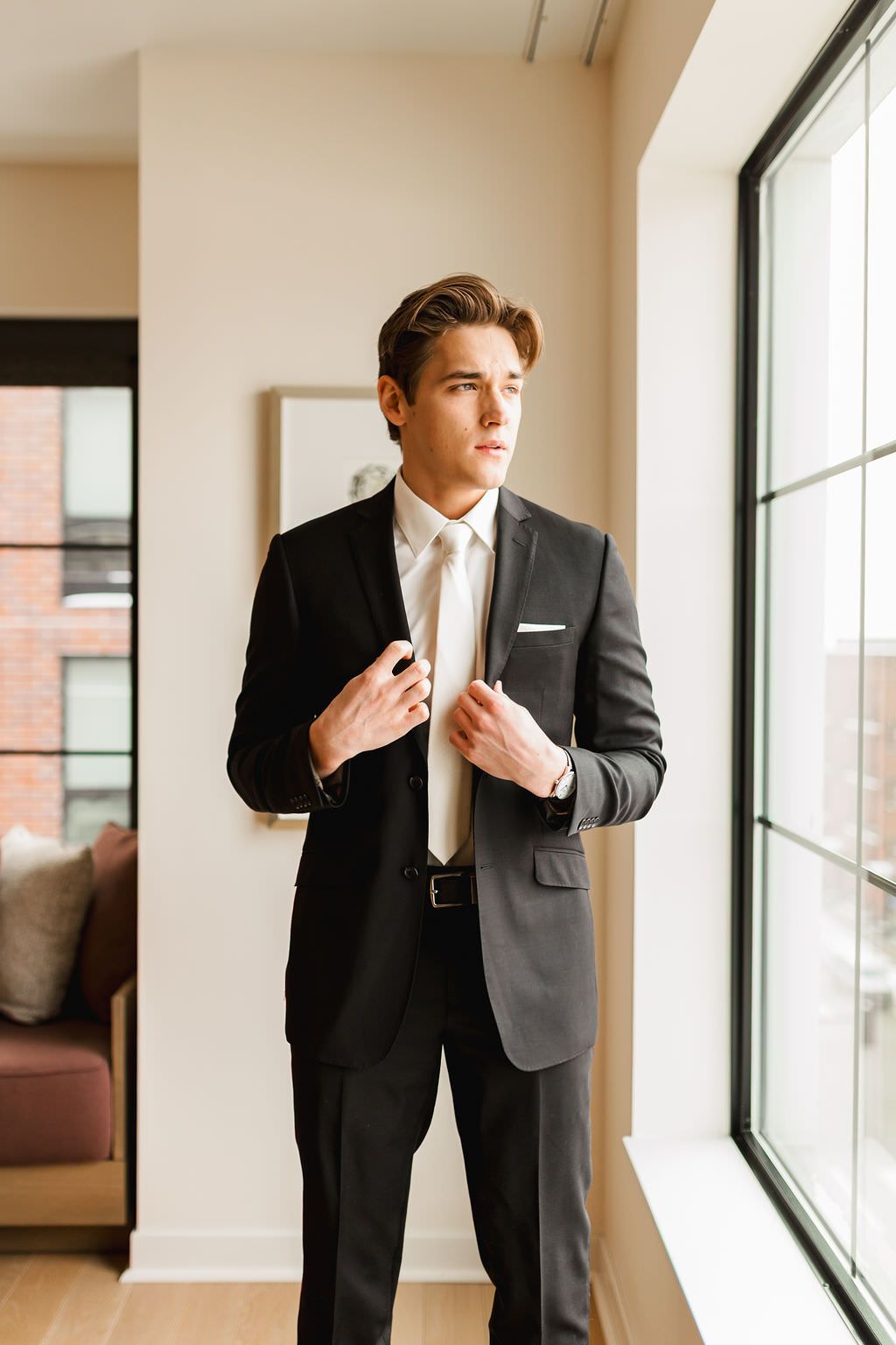 A groom adjusts his tie while standing in a bright room with large windows.