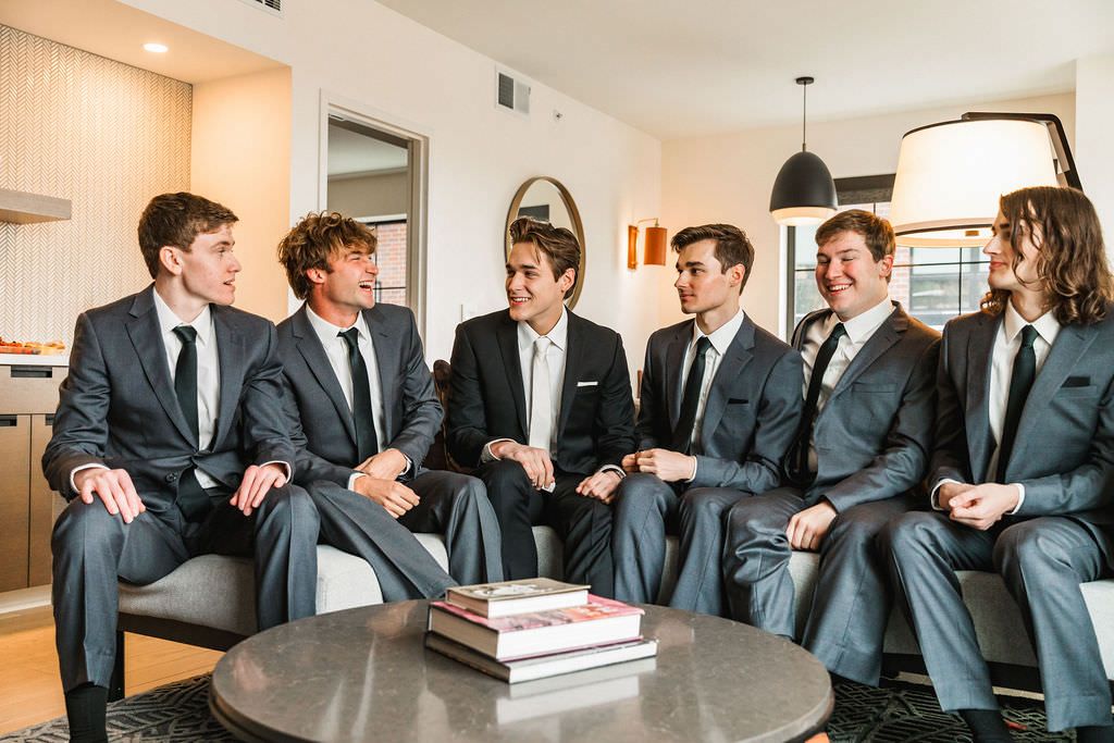 Groomsmen in dark suits stand together joking and laughing in a bright indoor hallway.