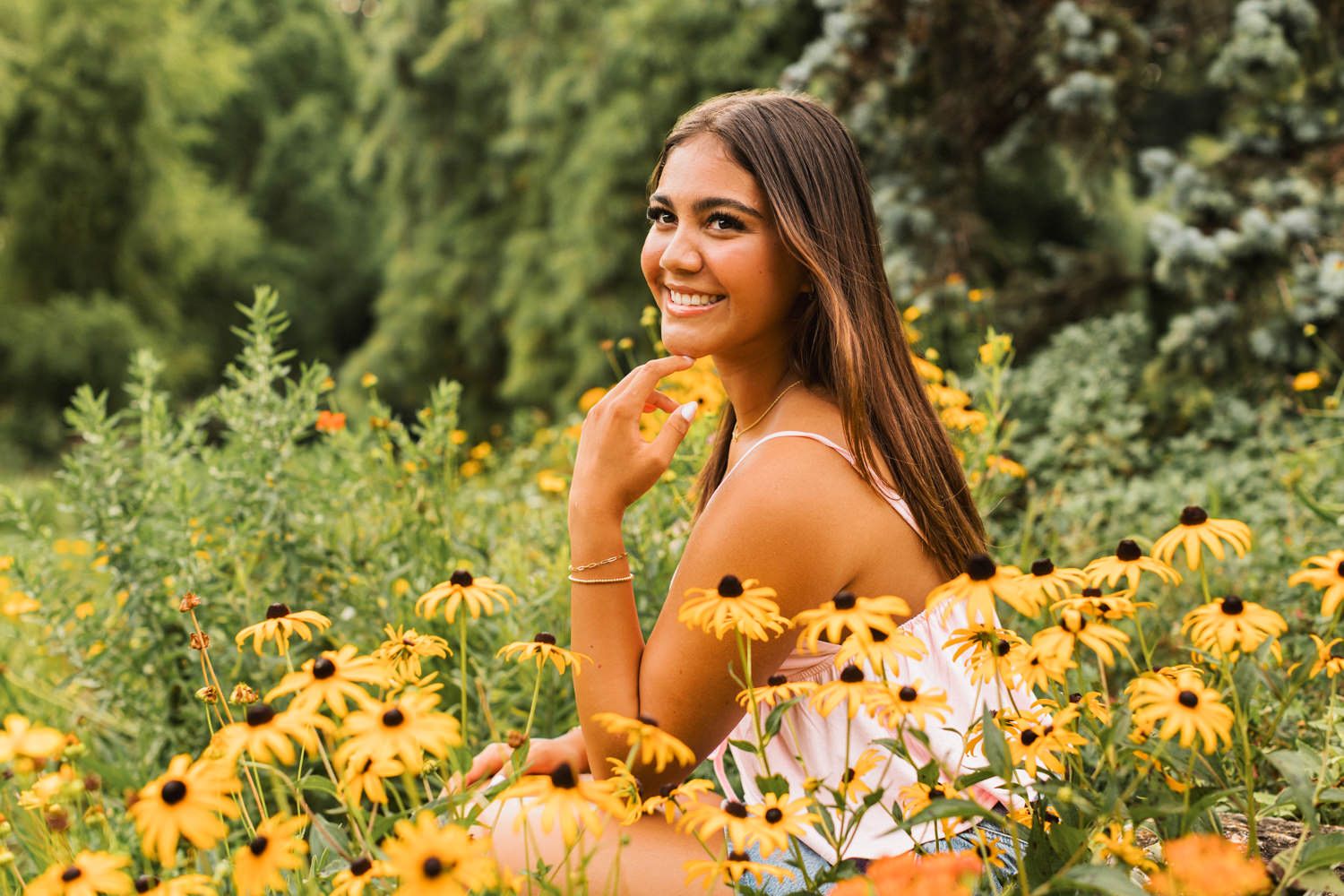 Someone poses in a lush garden setting surrounded by vibrant yellow flowers and greenery on a sunny day.