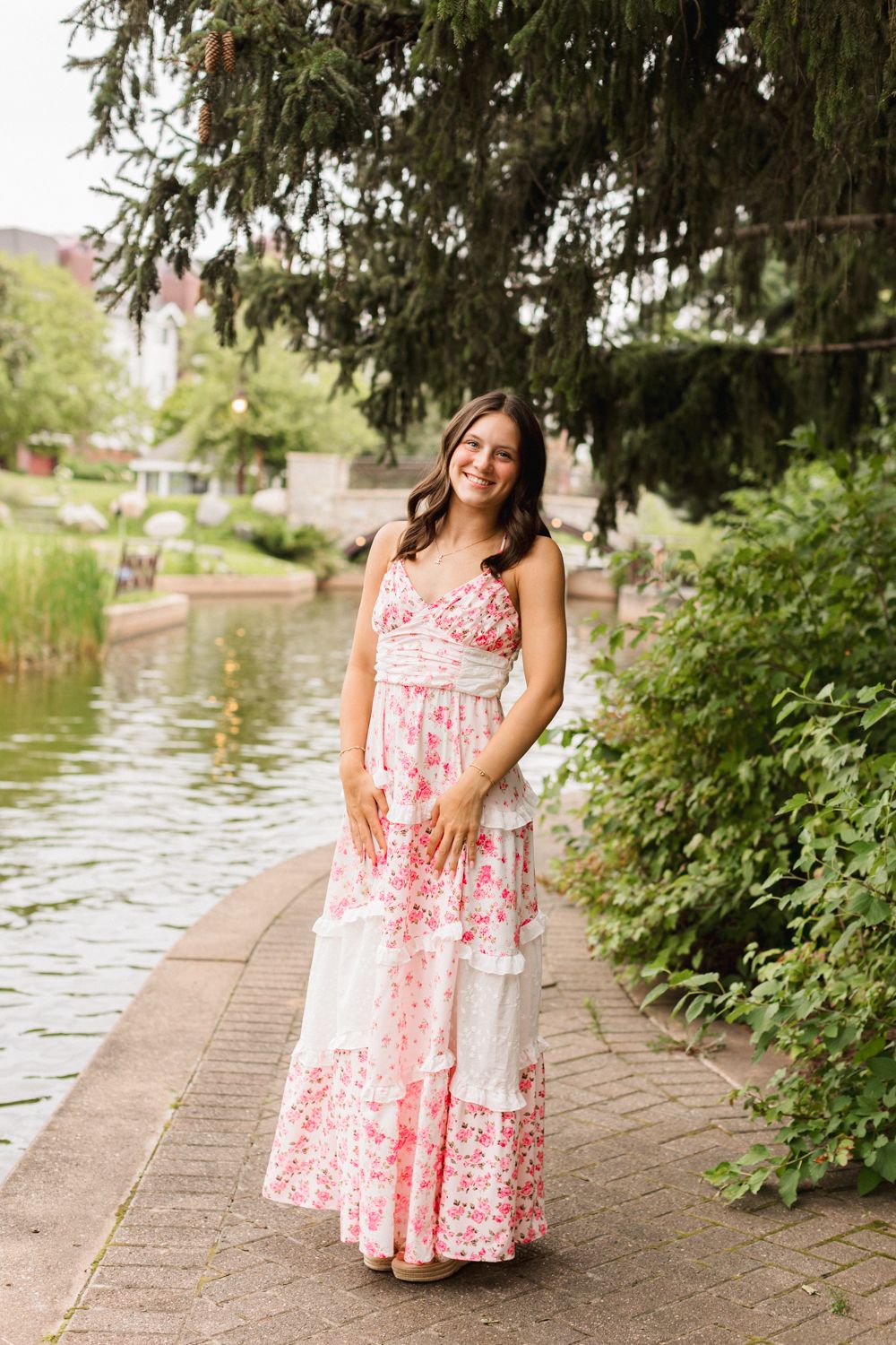 A person in a pink and white floral sundress poses along a stone pathway next to a serene pond on a summer day.