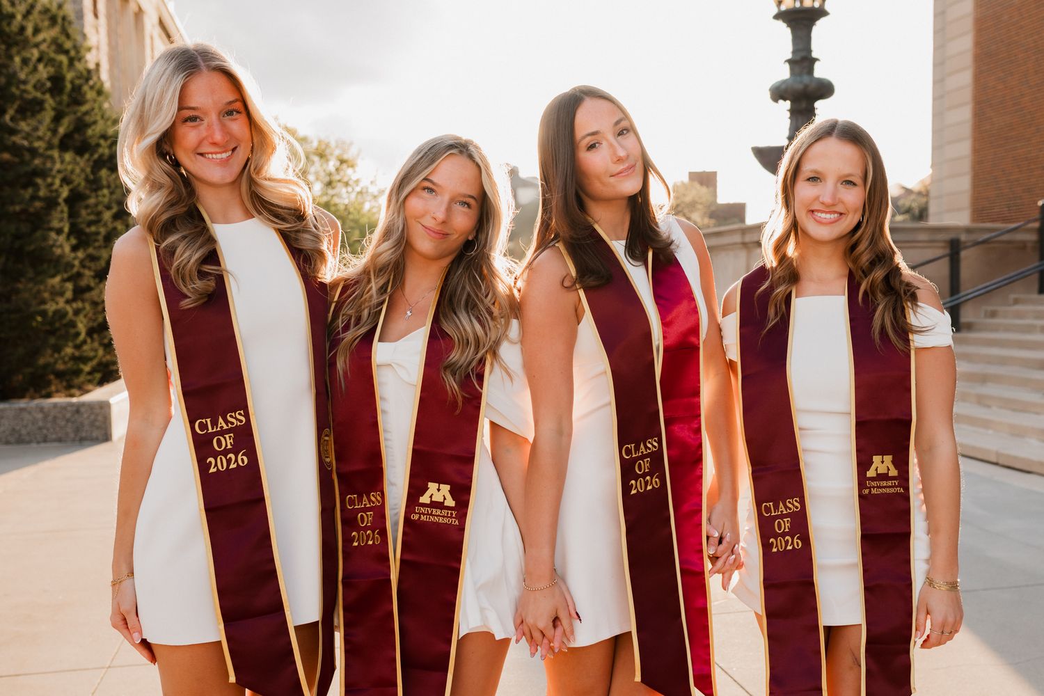 Four smiling University of Minnesota Class of 2024 graduates posing in white dresses with maroon graduation stoles.