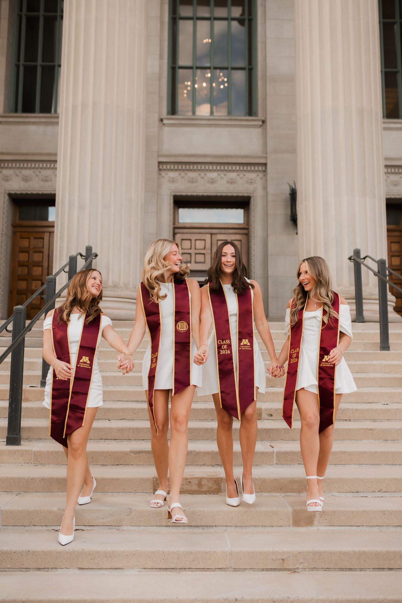 Four female graduates in white dresses and maroon graduation stoles walk down stone steps of a university building.