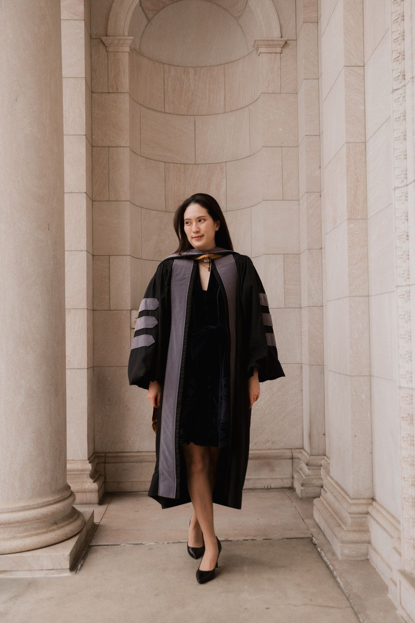 A woman in a black doctoral graduation gown poses between elegant stone columns at a university building.