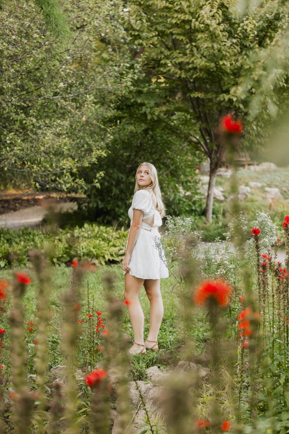 A person in a white dress walks through a lush garden with vibrant red poppies and green foliage.