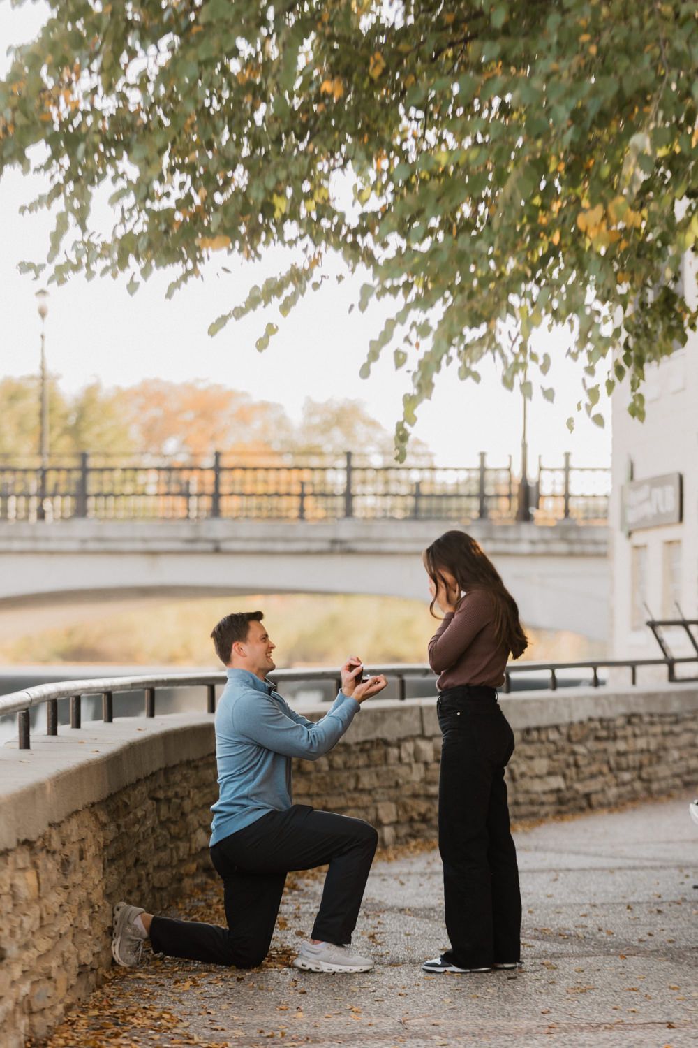 A romantic proposal scene on a stone pathway near a bridge during autumn with falling leaves overhead.