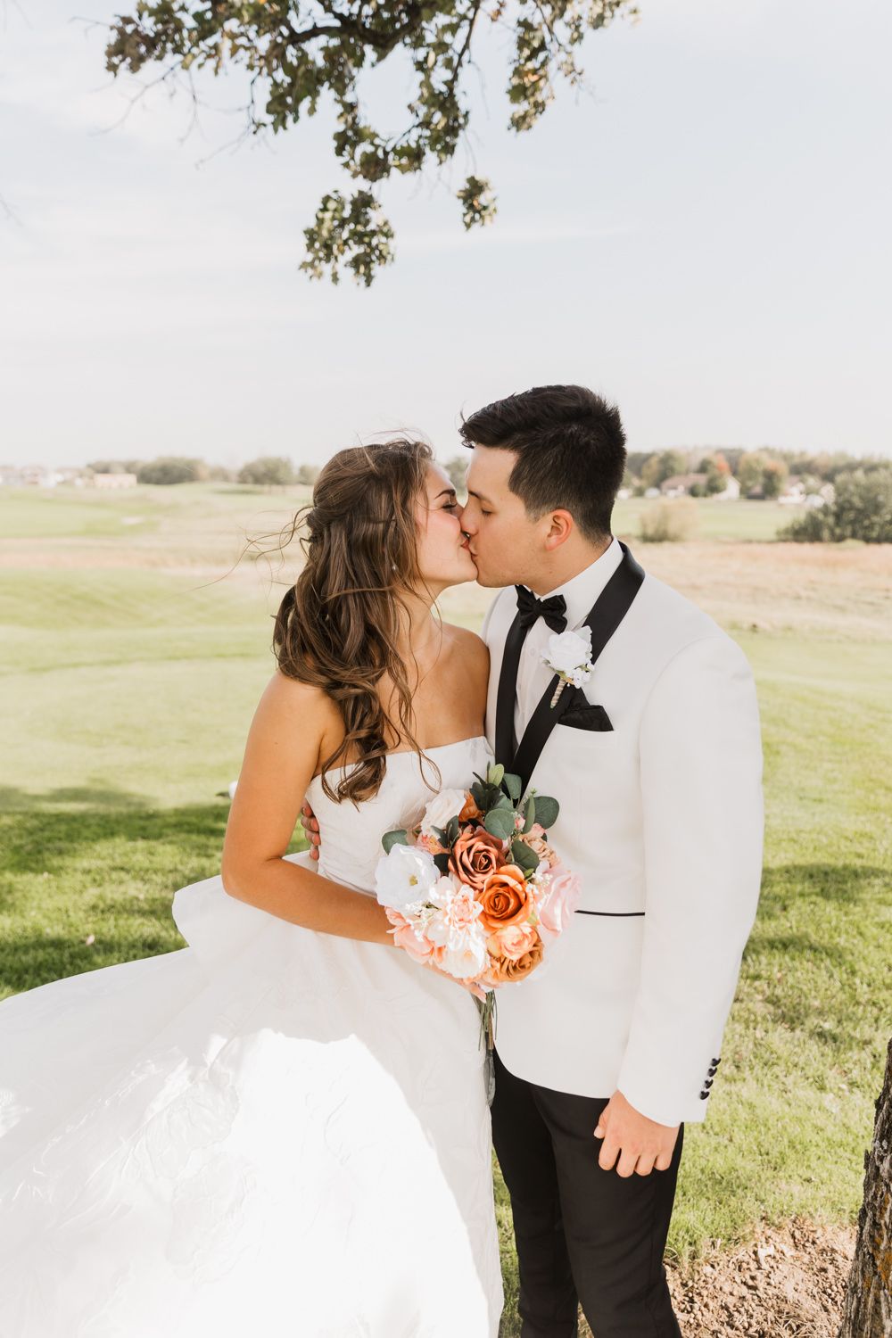 Newlywed couple shares intimate moments in a sunlit field with peach bouquet and elegant attire.