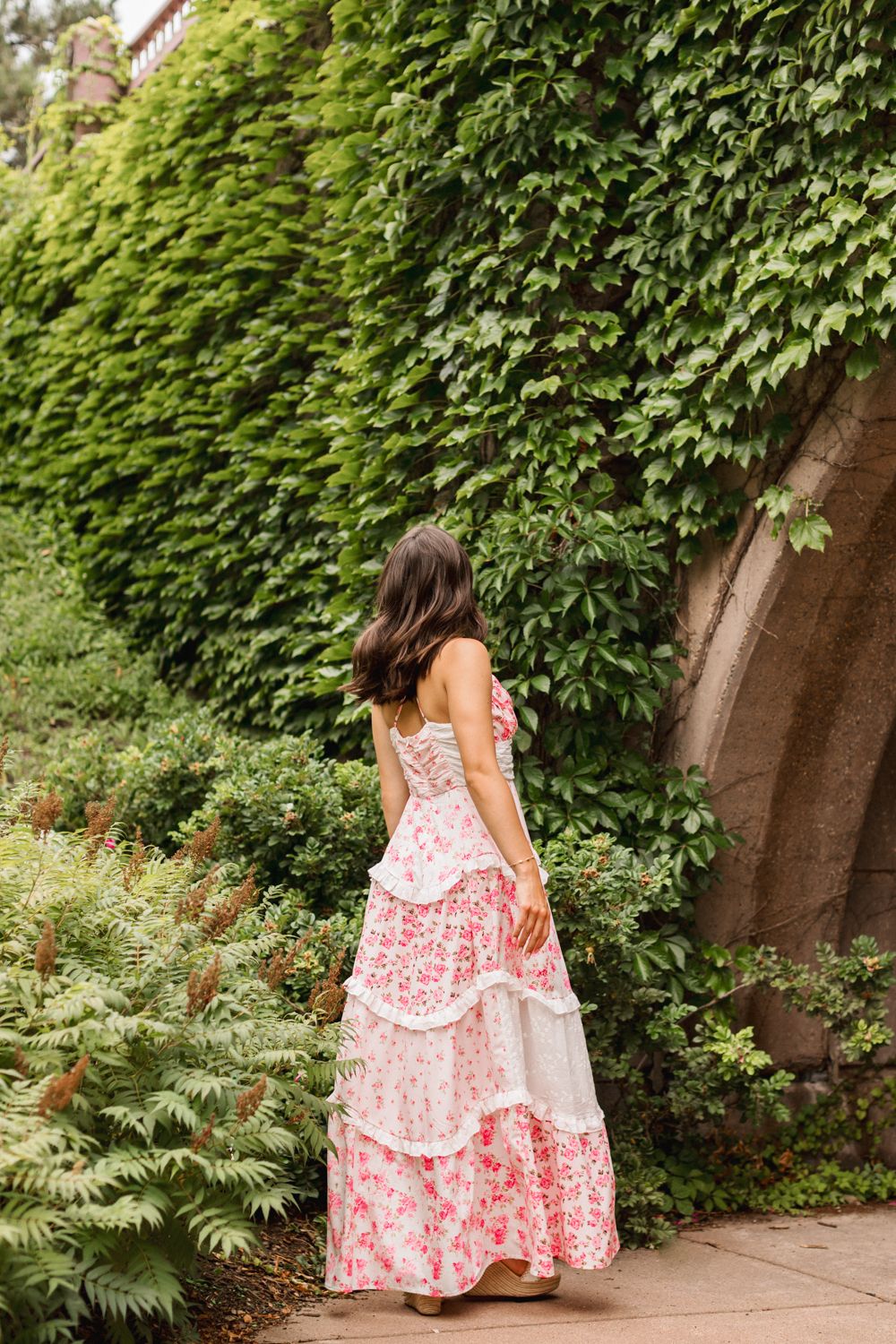 A woman in a pink and white floral maxi dress stands in front of an ivy-covered archway in a garden setting.
