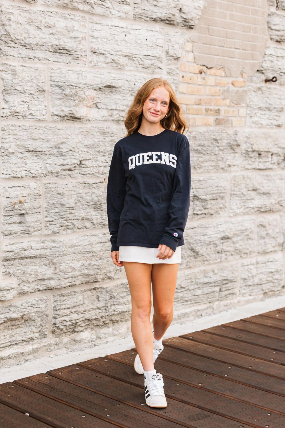 Person in navy blue college sweatshirt and white shorts poses casually on wooden deck against stone wall.