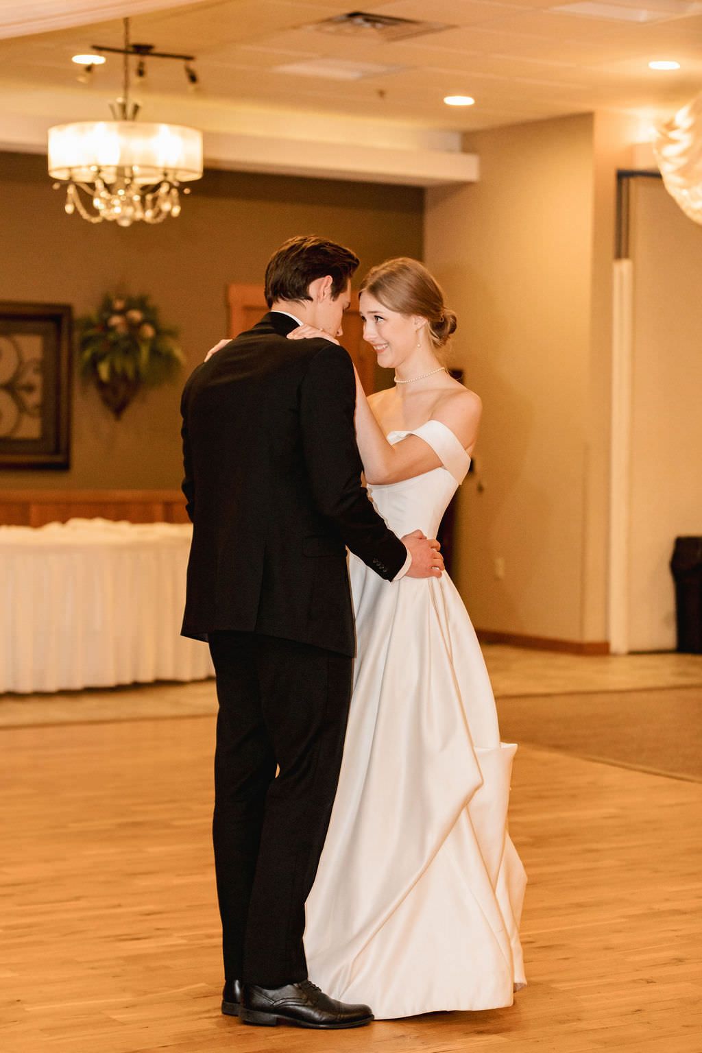 Newlyweds dancing together on wooden floor under chandelier lighting.