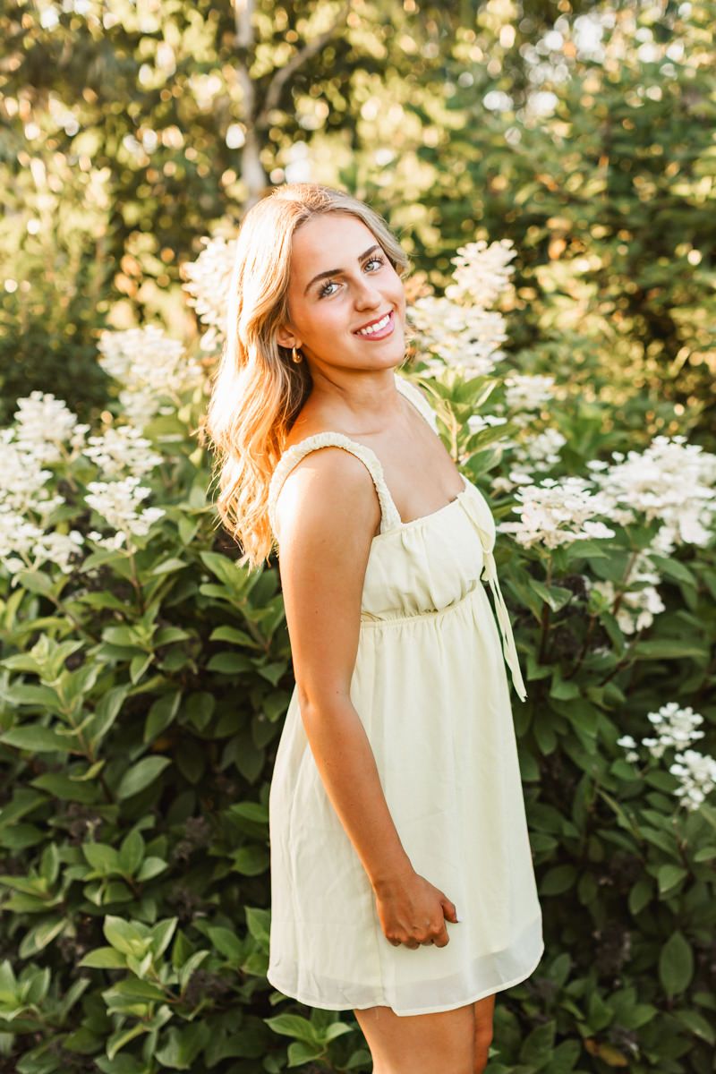 A person in a light yellow ruffled top poses outdoors against greenery and white flowers in golden sunlight.
