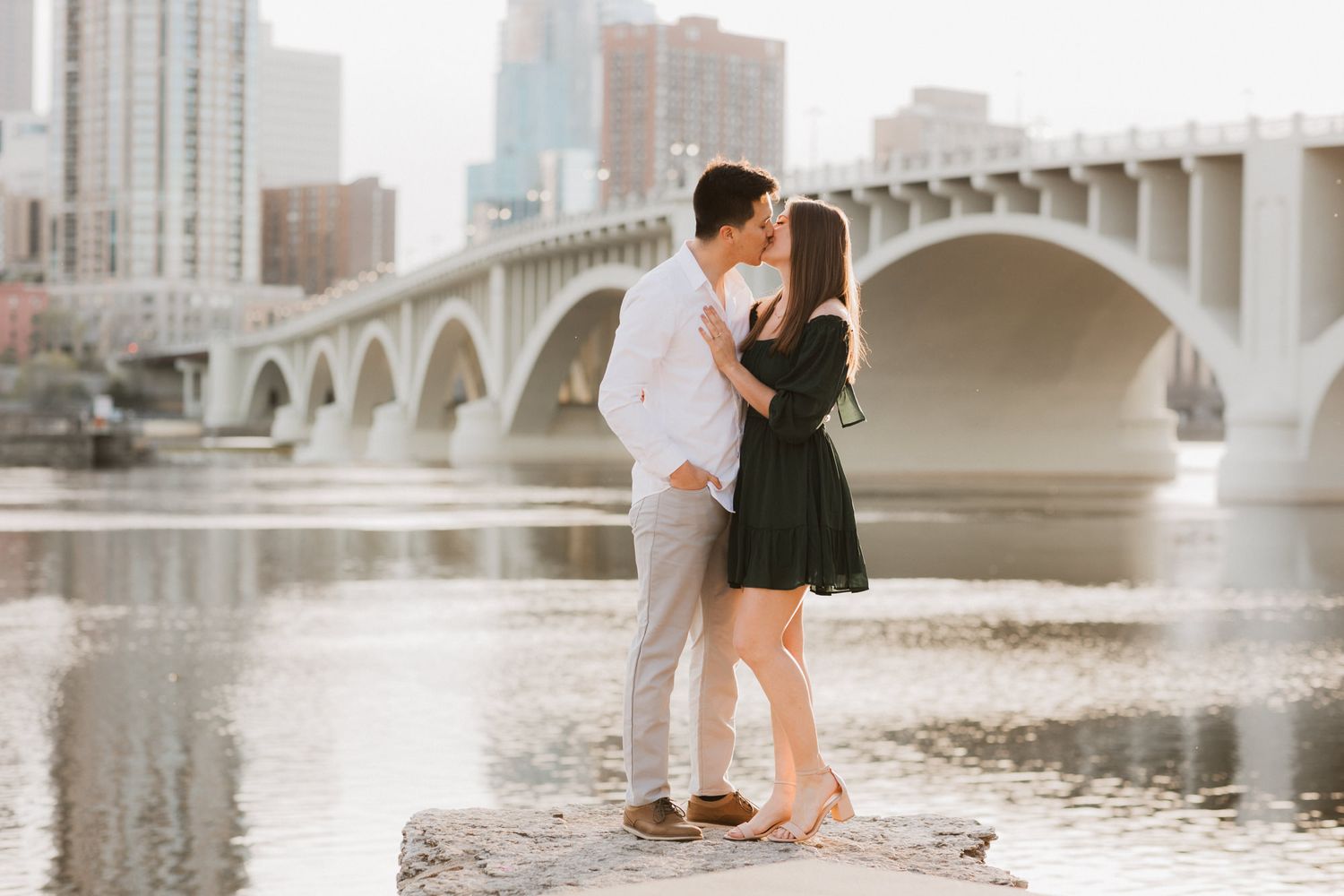 Couple kissing on rocks by a river with a bridge and city skyline in the background.