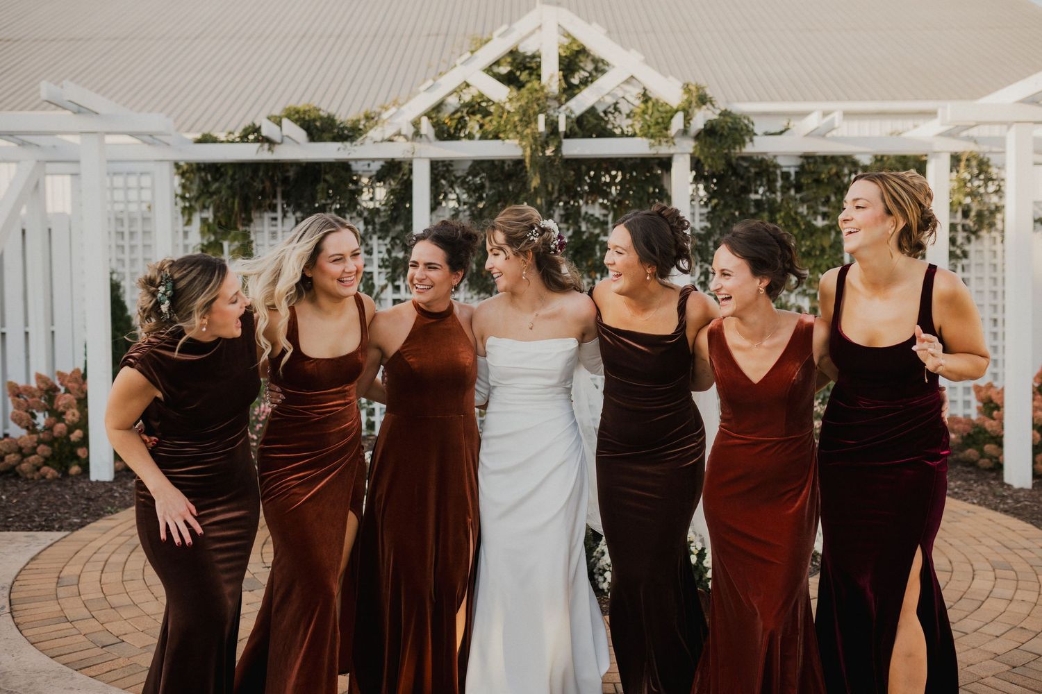 A laughing bride in a white gown stands arm in arm with her bridesmaids in burgundy and brown velvet dresses on a brick patio.