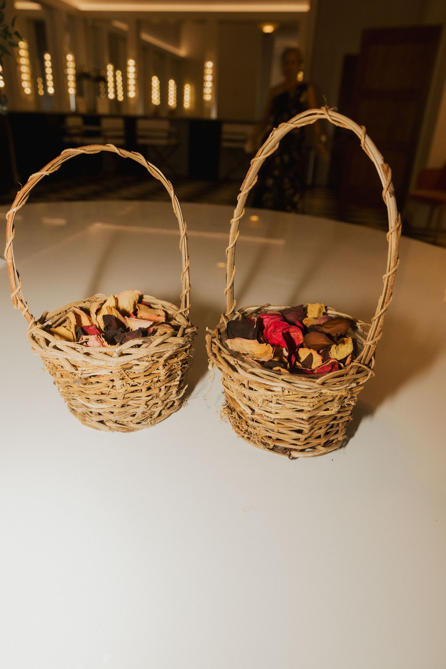 Two small woven wicker baskets with handles filled with dried flower petals sit on a white table at a wedding venue.