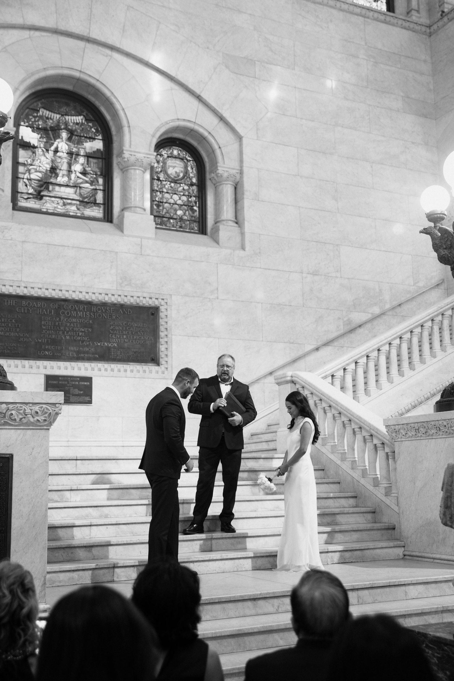 Black and white photo of a bride and groom exchanging vows on grand staircase with officiant at elegant wedding ceremony.