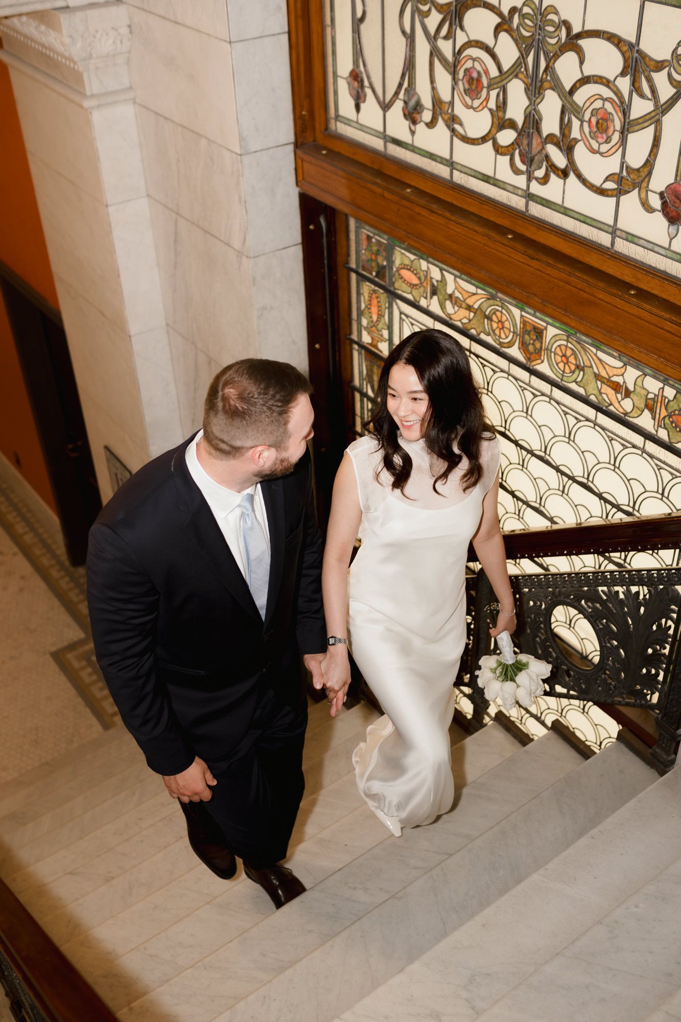 Bride and groom walking up ornate staircase, bride in white gown holding bouquet, groom in black suit.