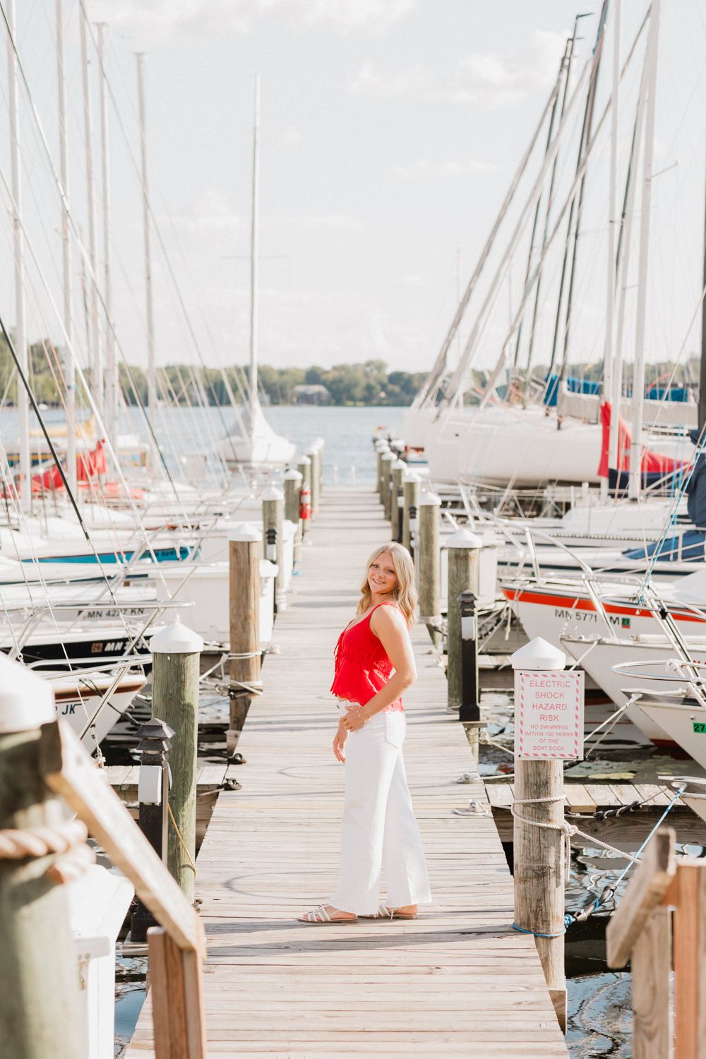 Woman in white dress walks along wooden dock with sailboats moored at marina on sunny day.
