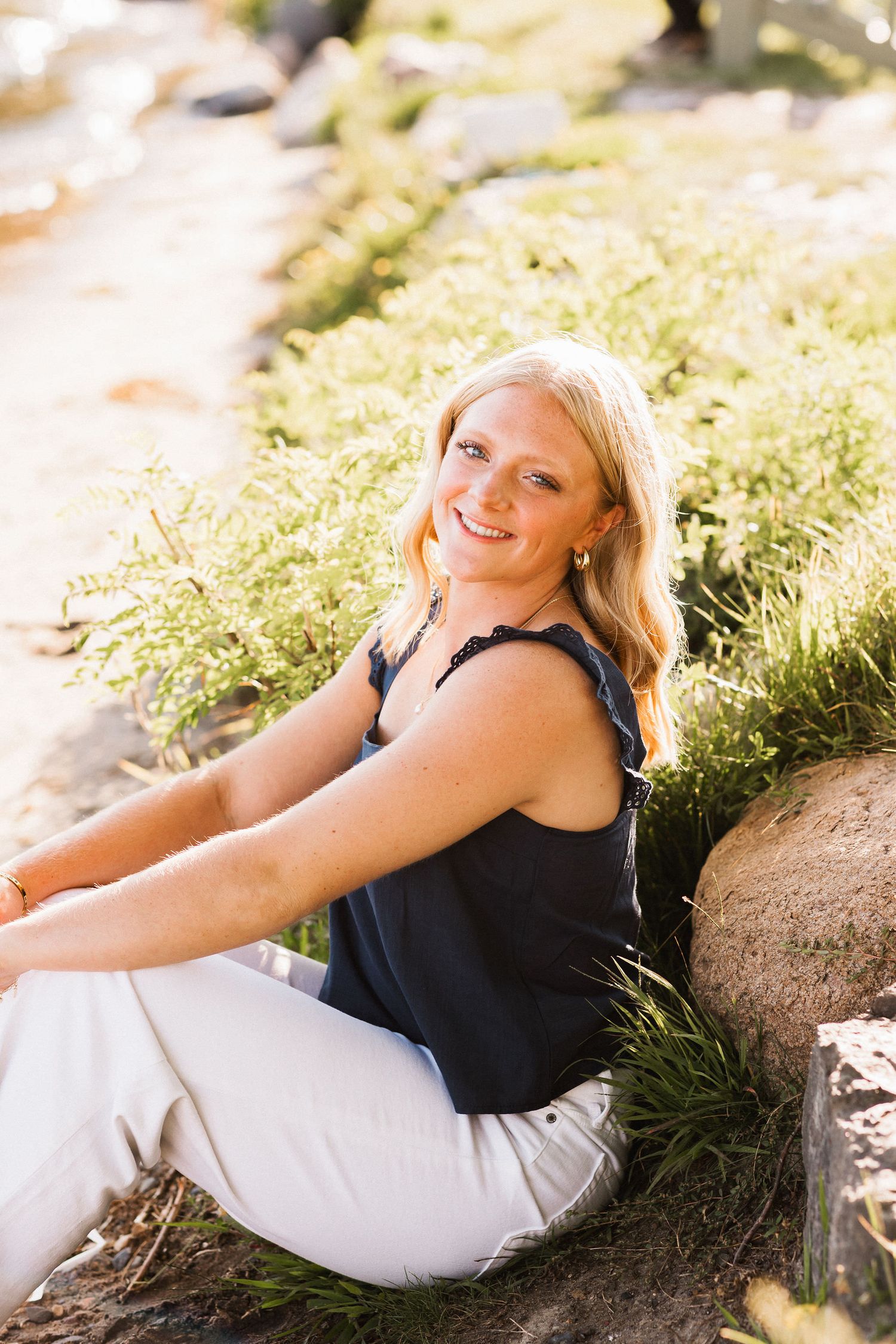 A person in black and white attire poses casually on a rock in dappled sunlight during an outdoor photo session.