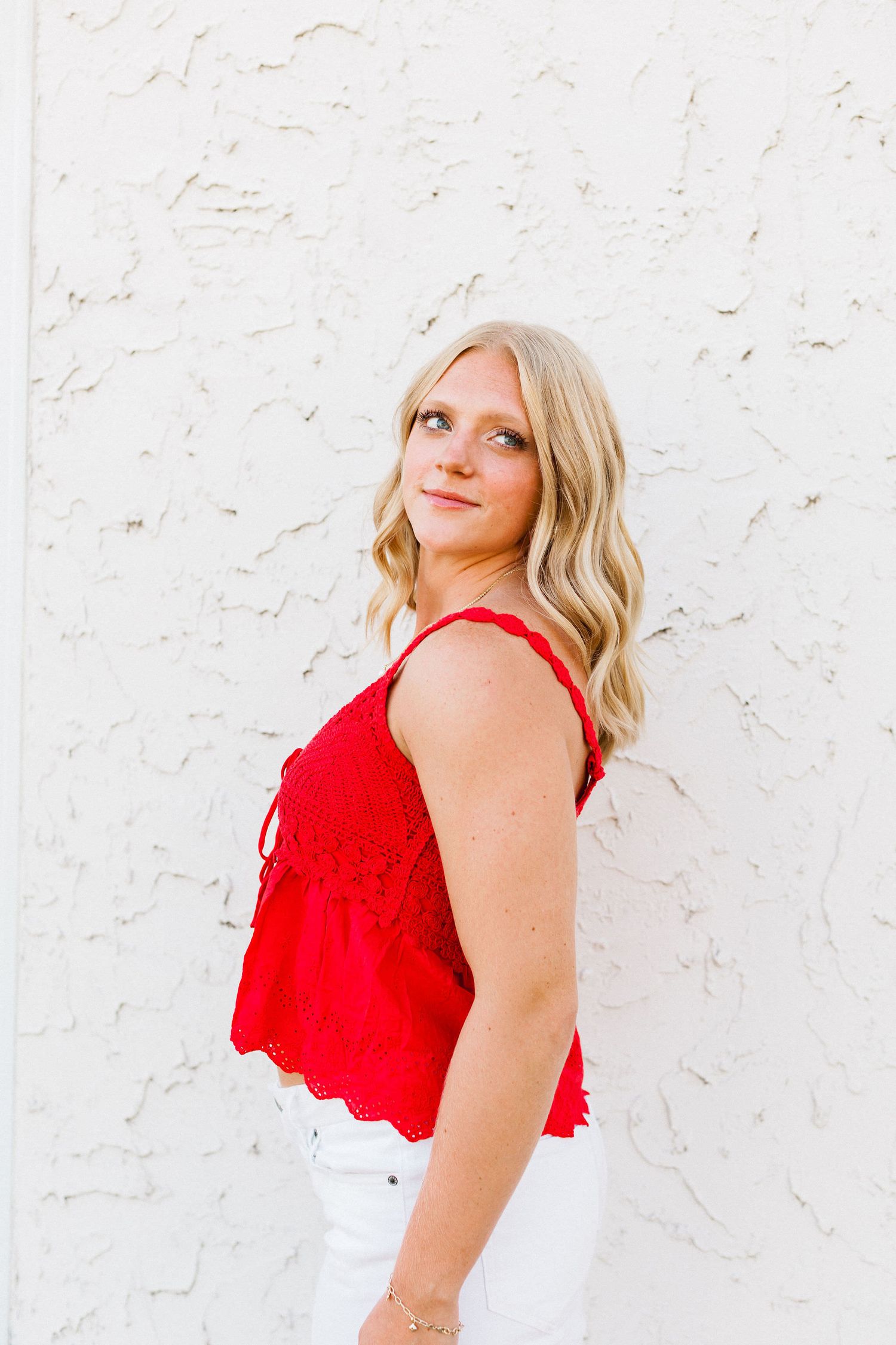 Woman in vibrant red sundress poses against white textured wall in summer photoshoot series.