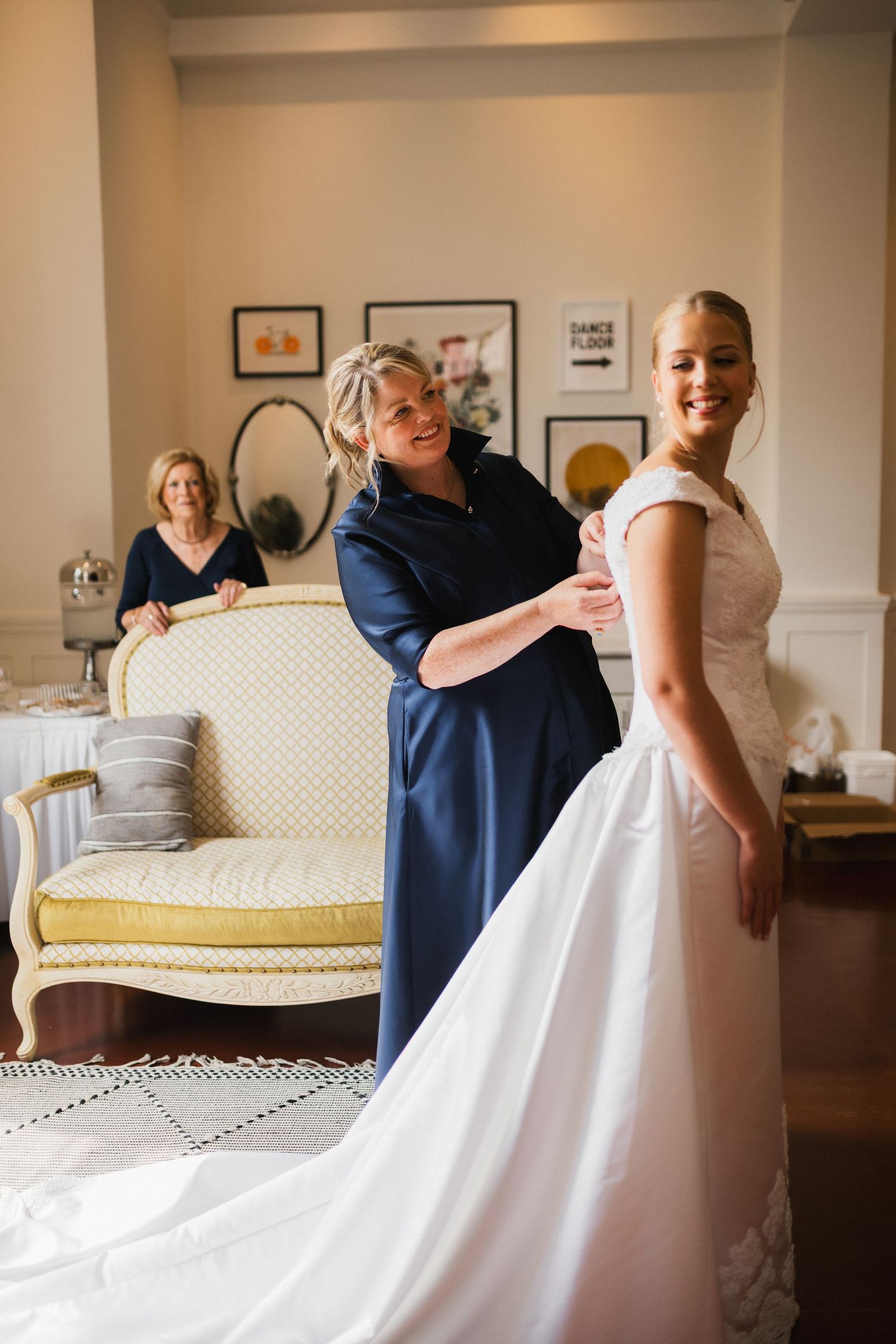 Bridesmaid in navy dress helping bride into white wedding gown while mother watches in elegant bridal suite.