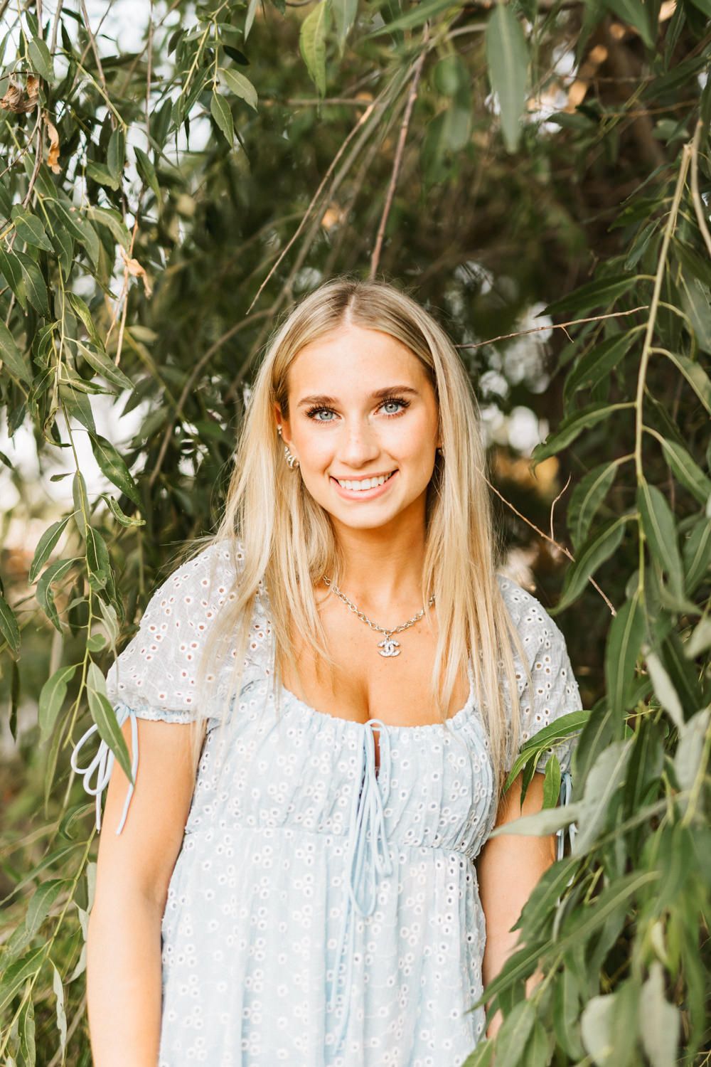 Woman in light blue dress poses among willow branches in natural outdoor setting.