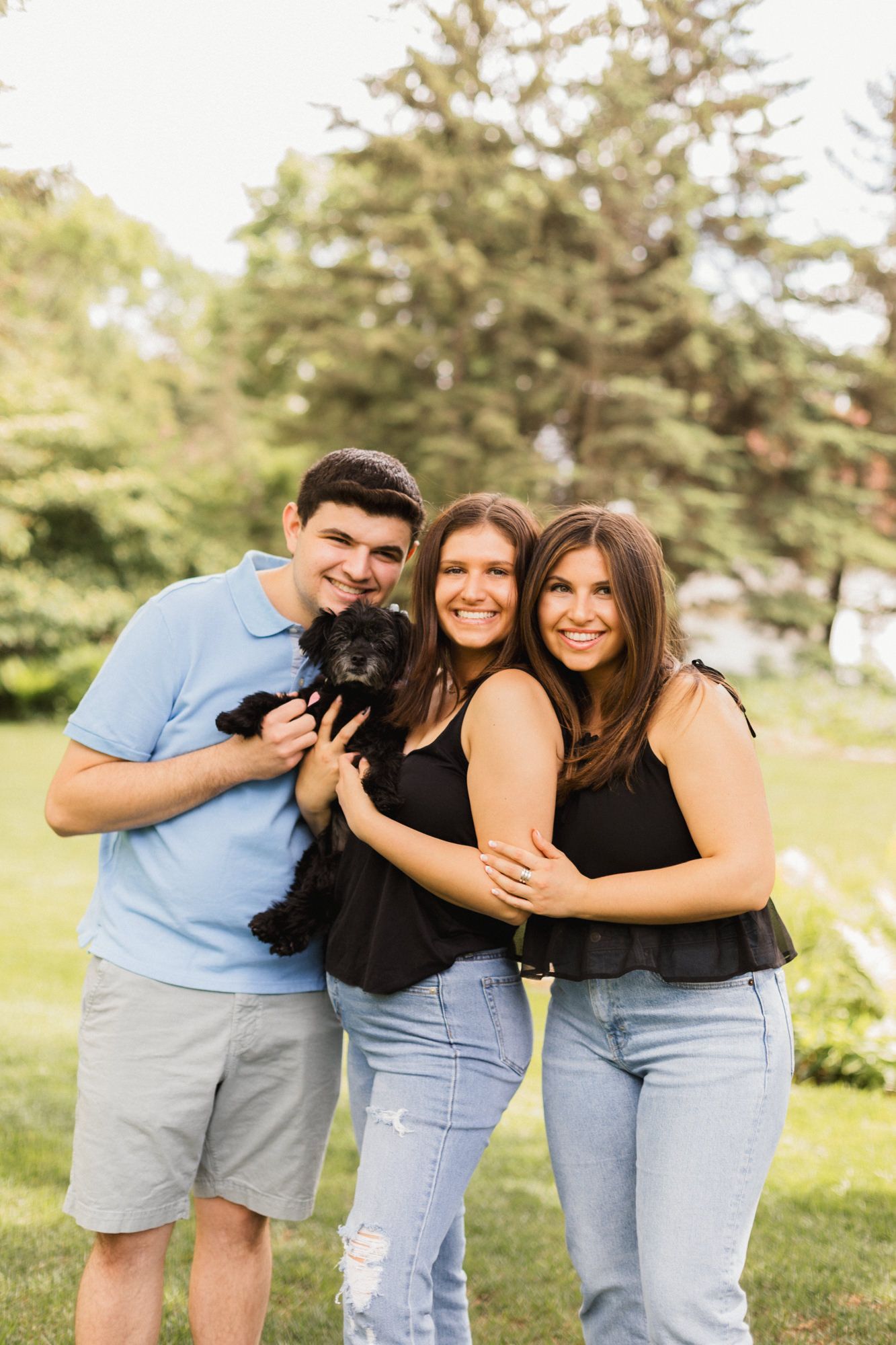 Three young adults smiling outdoors holding a small black dog in a sunny green park.