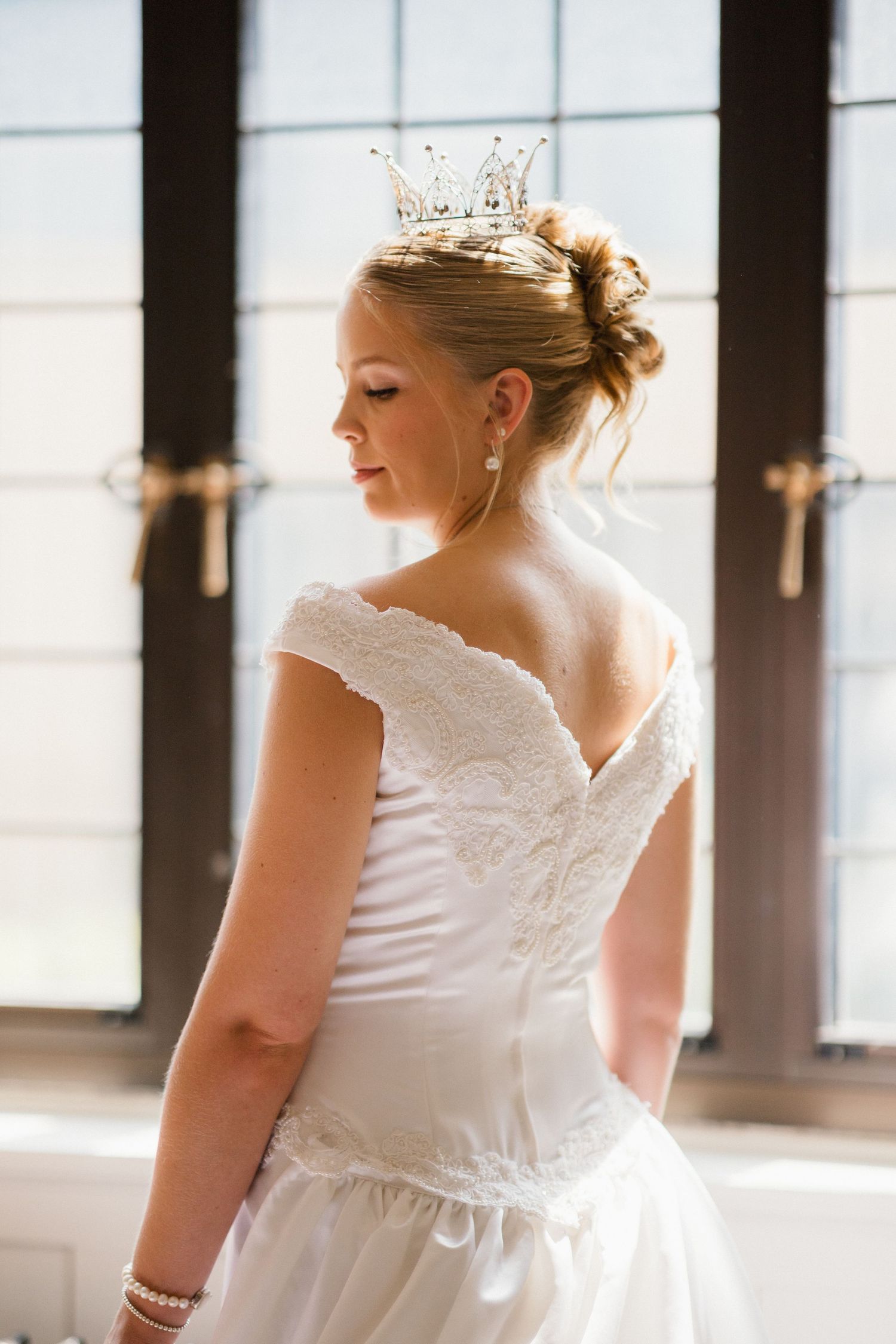 Bride in white lace wedding dress and crystal crown poses gracefully with back to camera near sunlit windows.