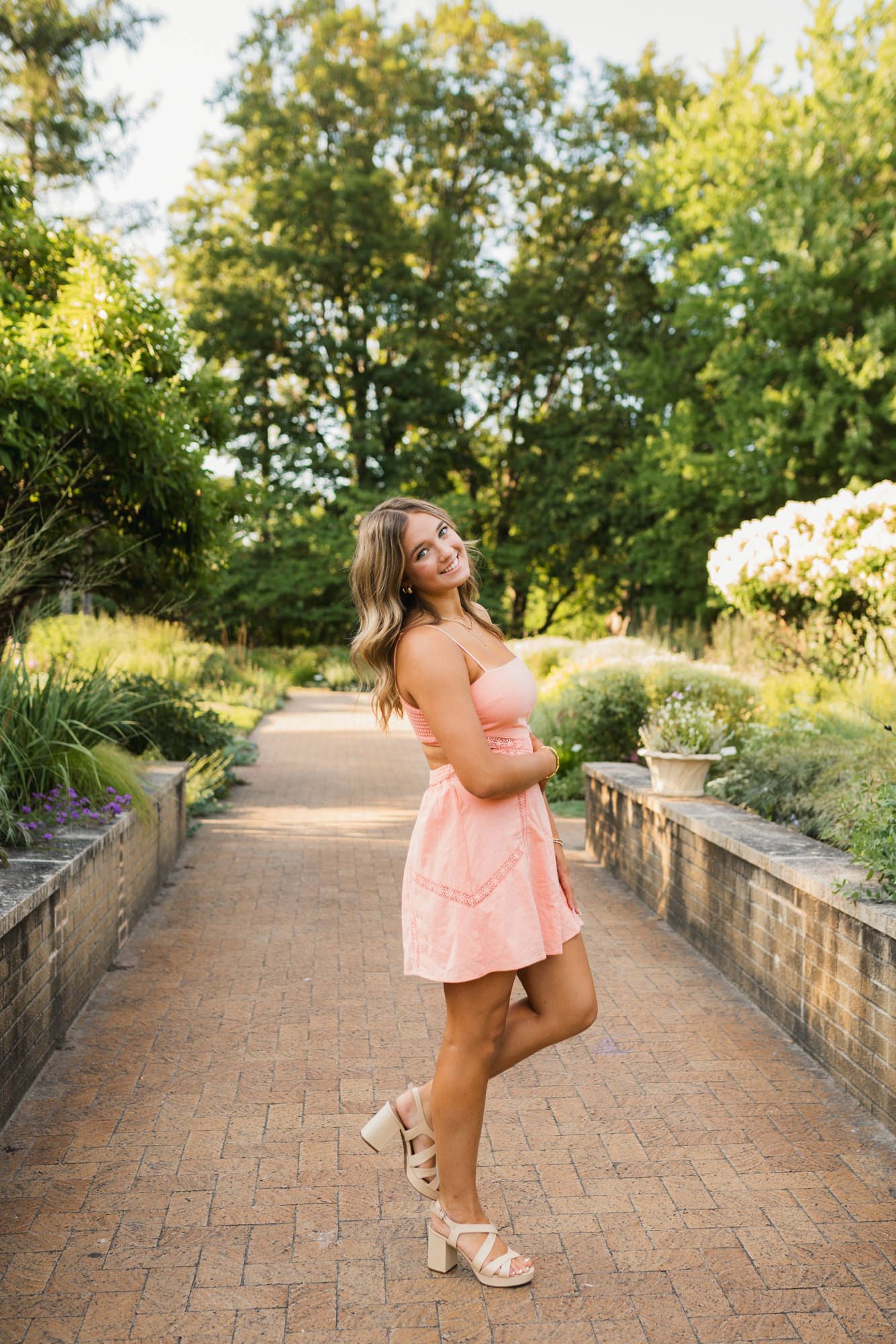 Young woman in pink mini dress and heels posing on brick garden path surrounded by lush greenery.