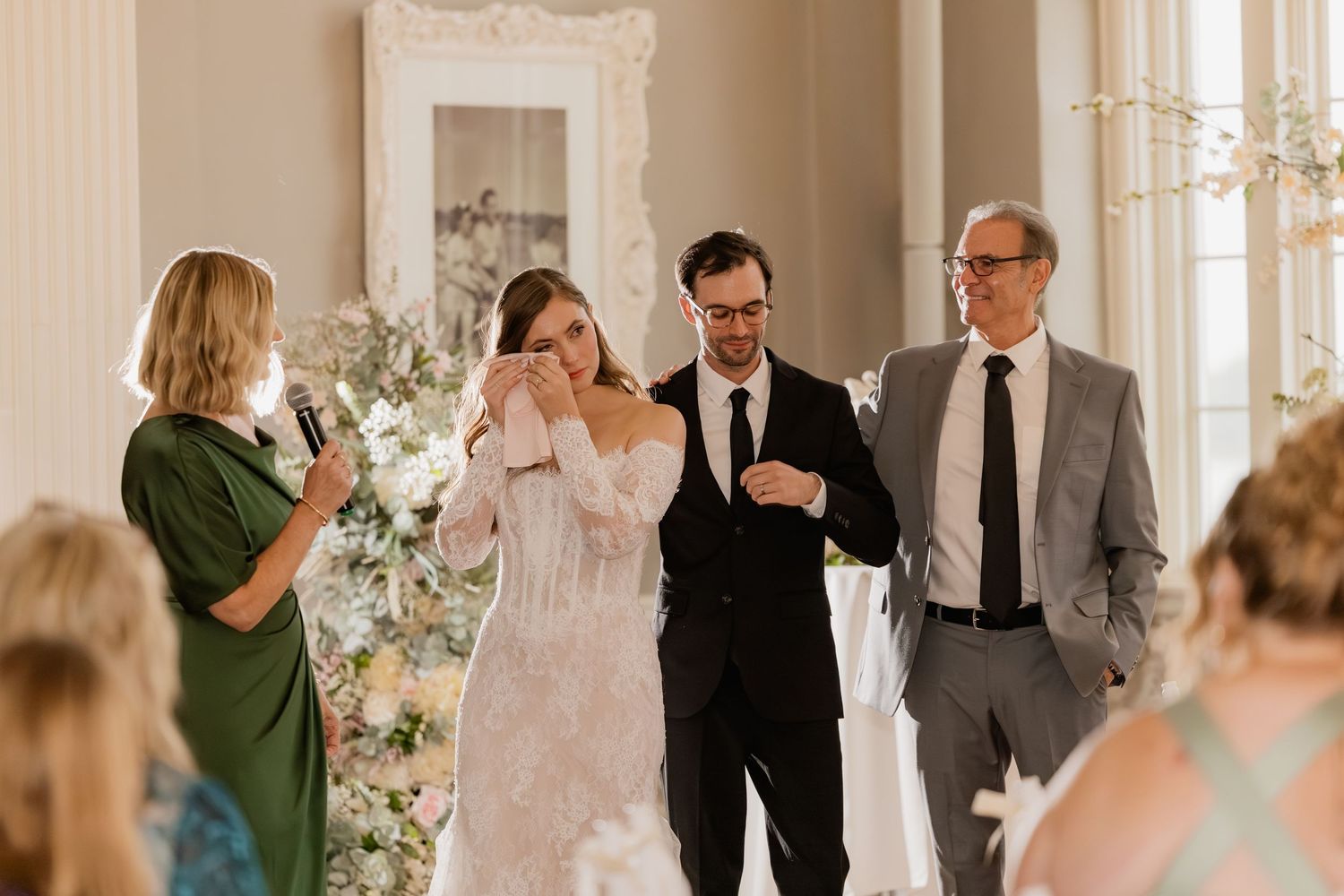 Emotional bride wiping tears with groom and father at indoor wedding ceremony with floral arrangements.